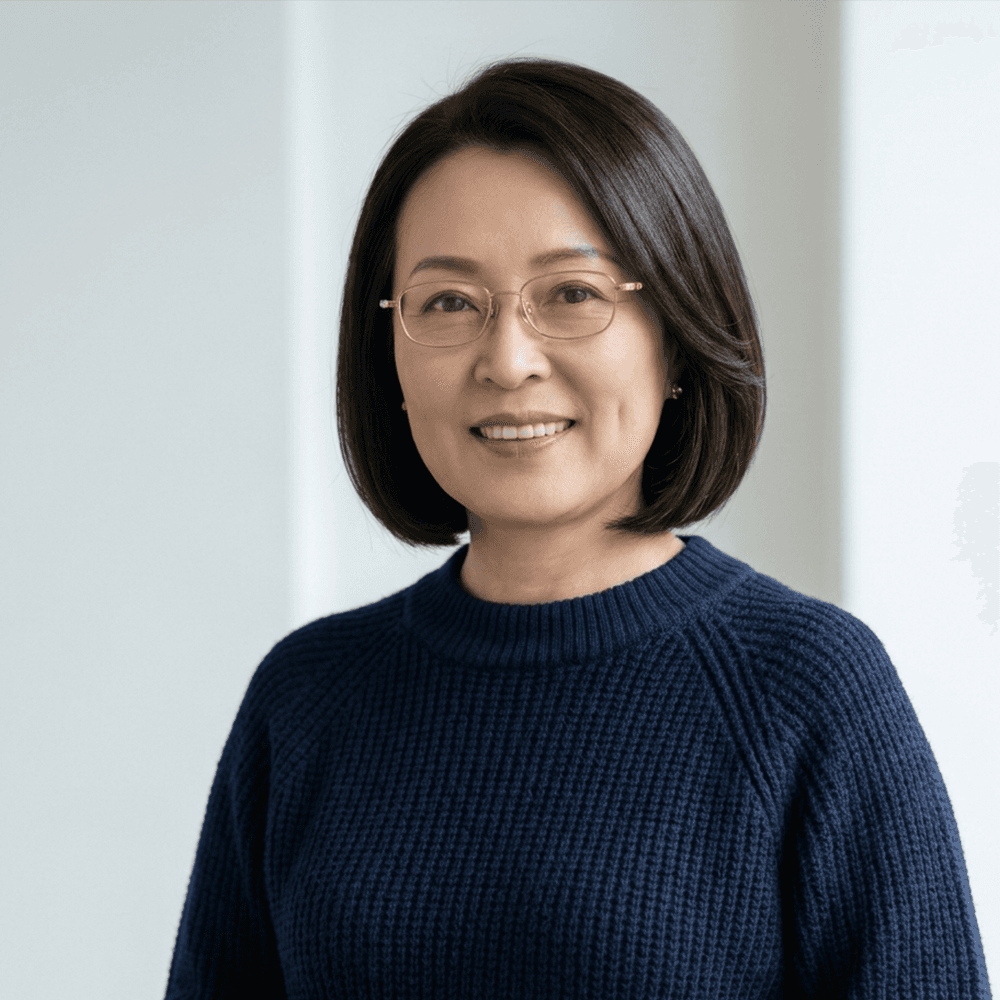 A medium close-up portrait photograph of an Asian woman in her late 40s or 50s with a dark bob haircut and gold-rimmed glasses, wearing a dark navy blue cable-knit sweater and smiling gently against a textured grey background.