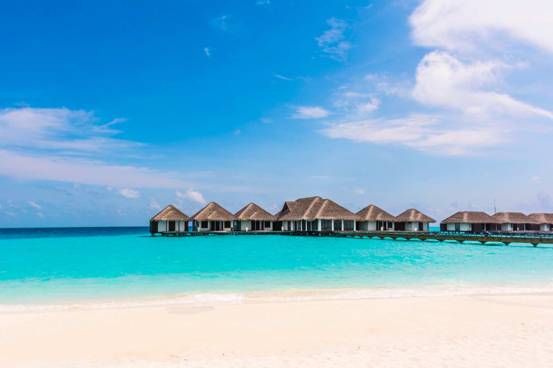 Overwater bungalows with thatched roofs sit in turquoise water along a white sand beach under a blue sky.