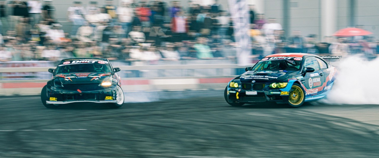 Two cars drift closely around a corner on a racetrack, surrounded by cheering spectators.