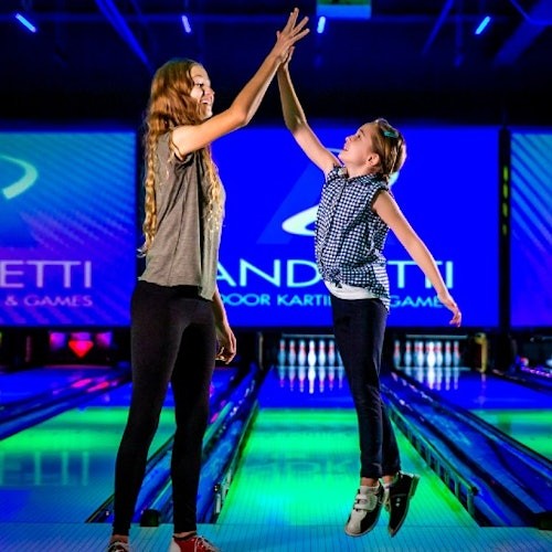 Двама души се поздравяват с високо петимата на оживена боулинг писта, като на екраните на заден план се вижда "Andretti Indoor Karting & Games".