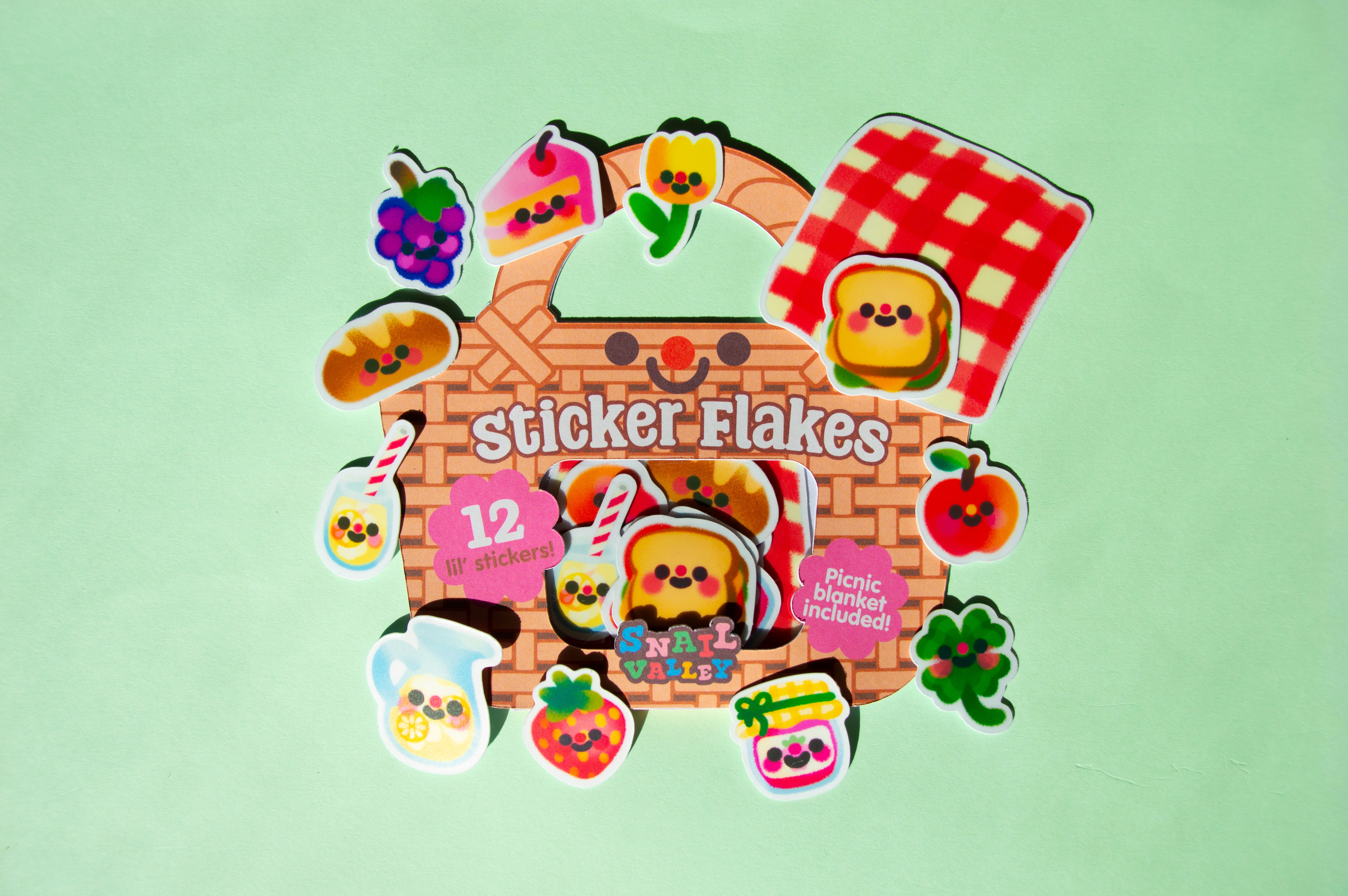 The picnic basket packaging on display with the small stickers included surrounding it. Some of the 12 stickers include: a sandwich, a picnic blanket, a strawberry, and a slice of cake.