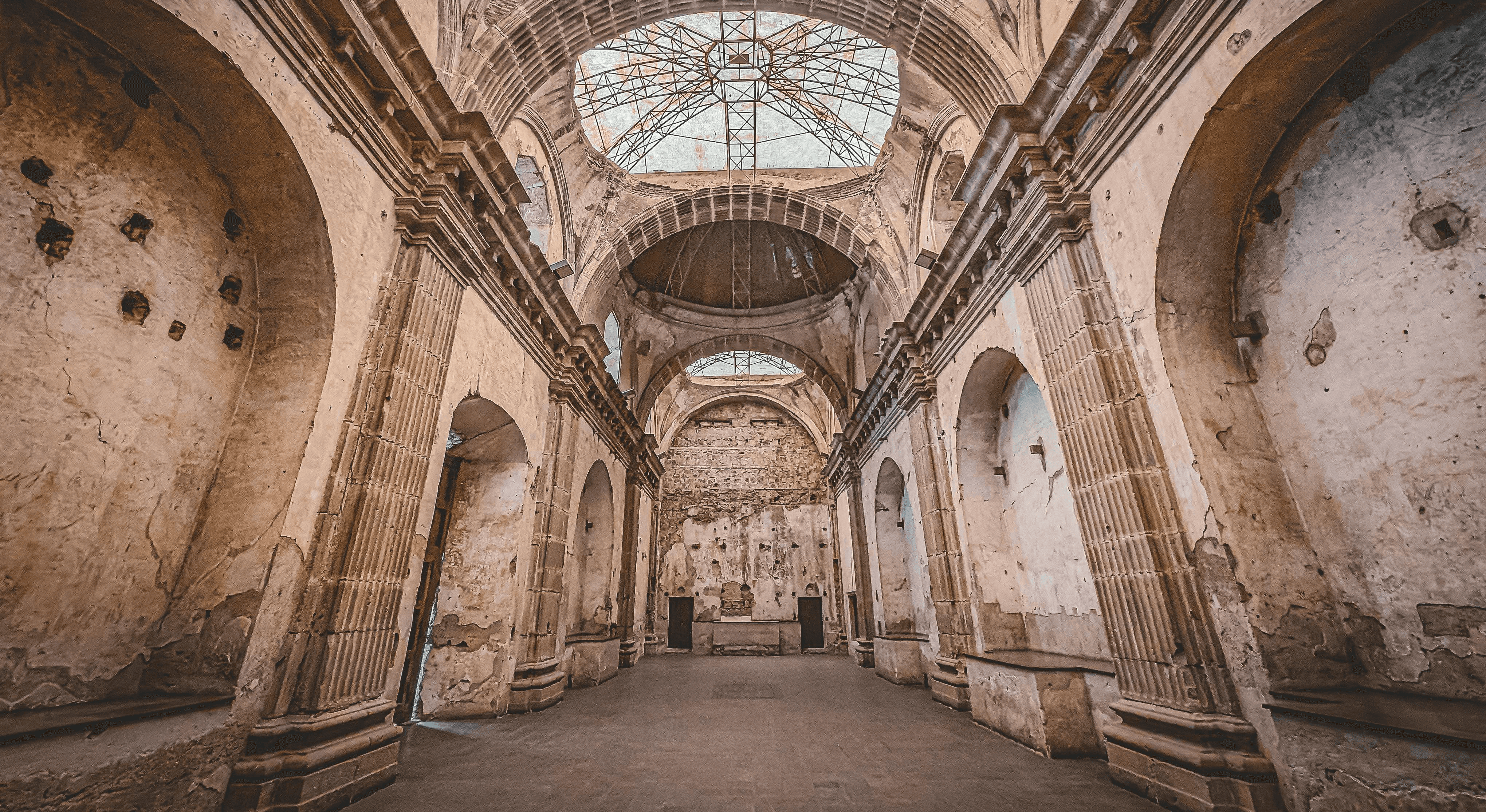 The image depicts the interior of an ancient, stone-arched cathedral with weathered walls, high ceilings, and a large, intricate dome with a partially open skylight, highlighting its historical architecture and religious heritage.