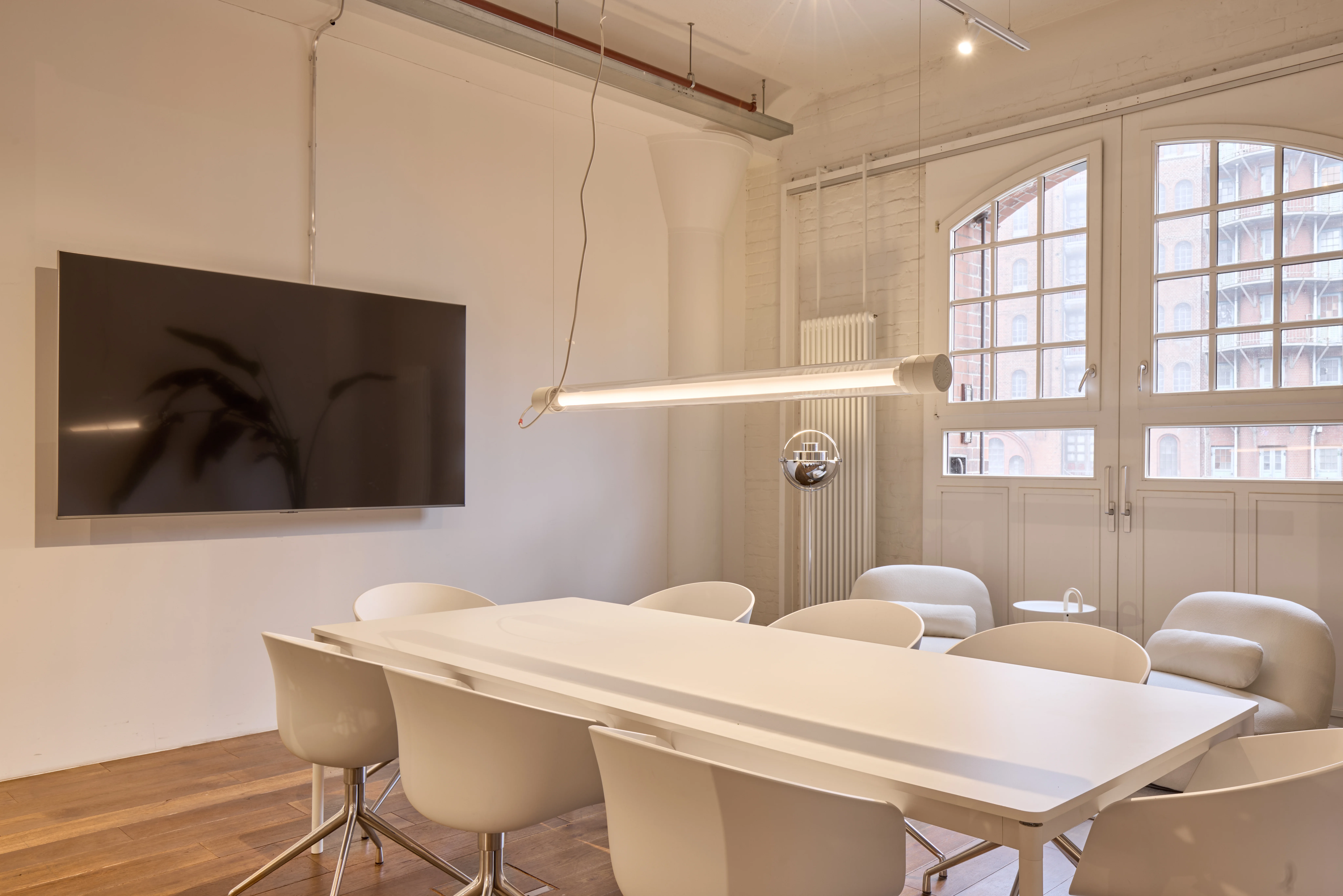 Bright conference room at INTERMATE OFFICE HAMBURG with a long white table, white shell chairs, a wall-mounted flat screen, a linear pendant light, and arched factory windows above white double doors.