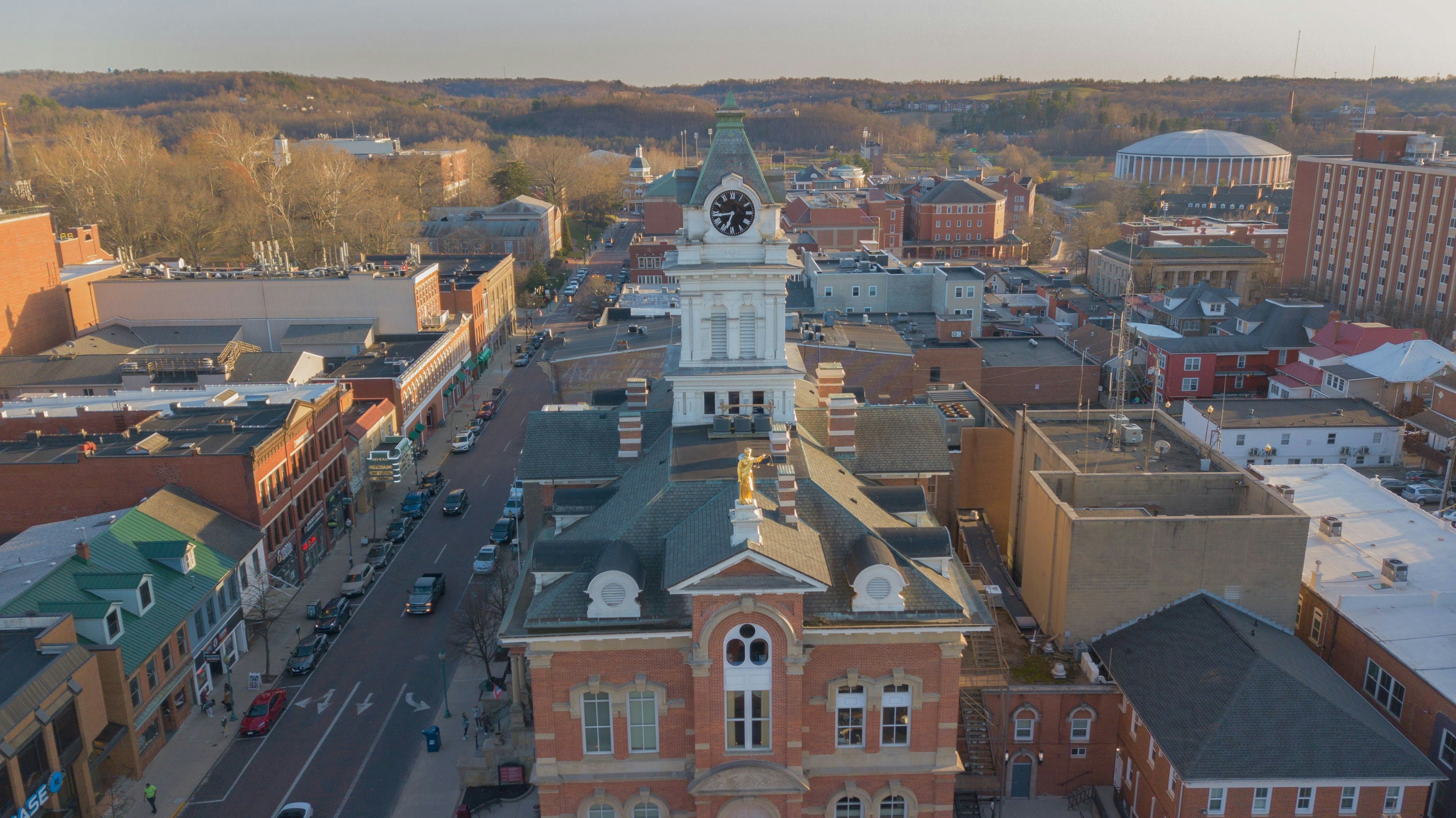 Aerial view of a historic brick courthouse in a Virginia city, complete with a clock tower. The surrounding streets are lined with historic buildings and cars, while rolling hills and trees are visible in the background, lit by the golden light of the setting sun.