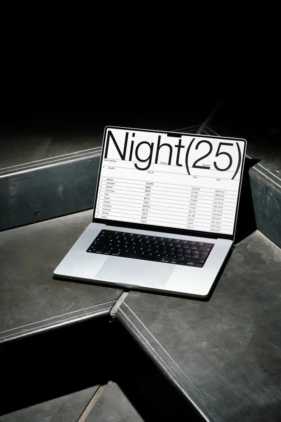 Open MacBook mockup placed on a dark, industrial surface, displaying a black screen in bold white typography and tabular data. Minimal lighting emphasizes the sleek, modern design.