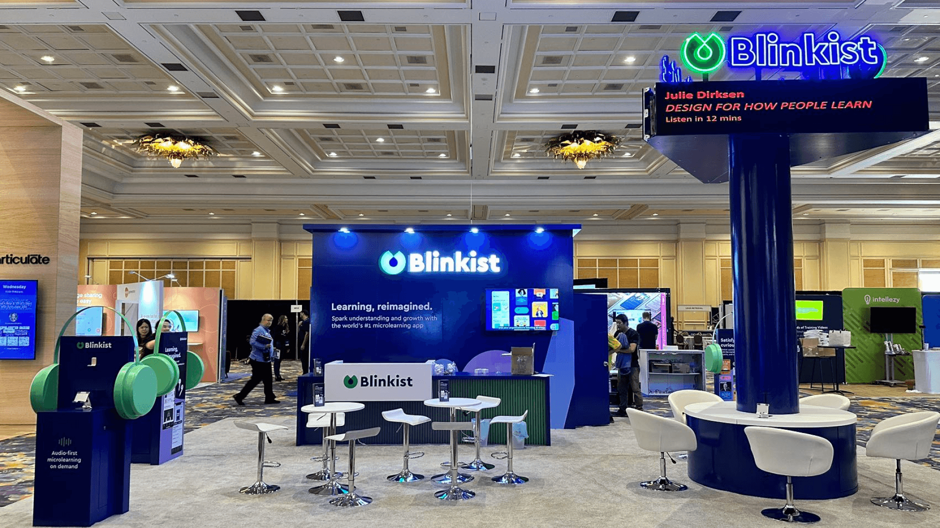 Blinkist DevLearn Conference & Expo 2025 20x40 technology product demo booth with branded back wall, reception counter, round meeting tables, headphones display, and central seating area