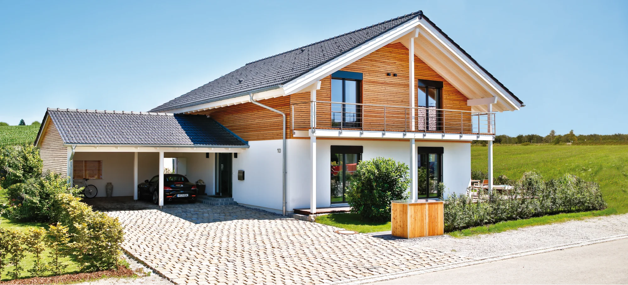 Modernes Einfamilienhaus mit Carport, Holzelementen und Balkon in ländlicher Umgebung unter blauem Himmel.