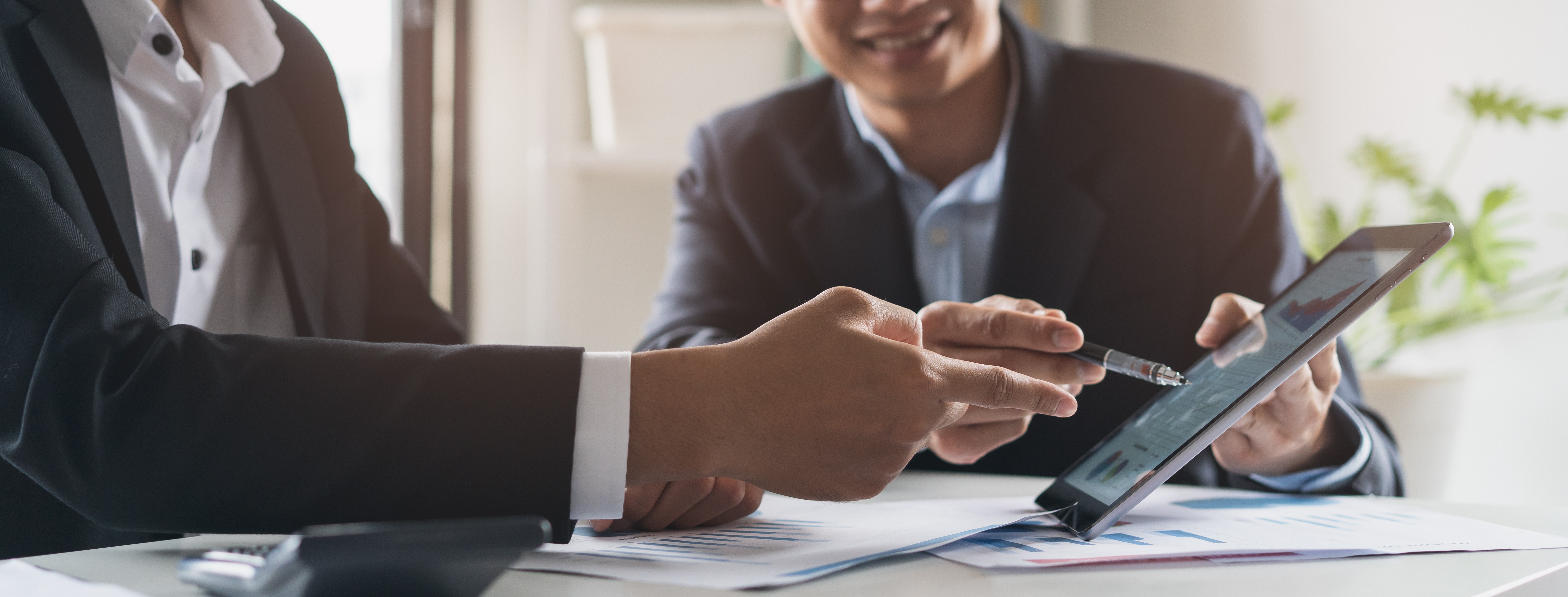 Zwei Geschäftsmänner im Anzug sitzen an einem Tisch und zeigen mit Stiften auf ein Tablet mit Diagrammen.