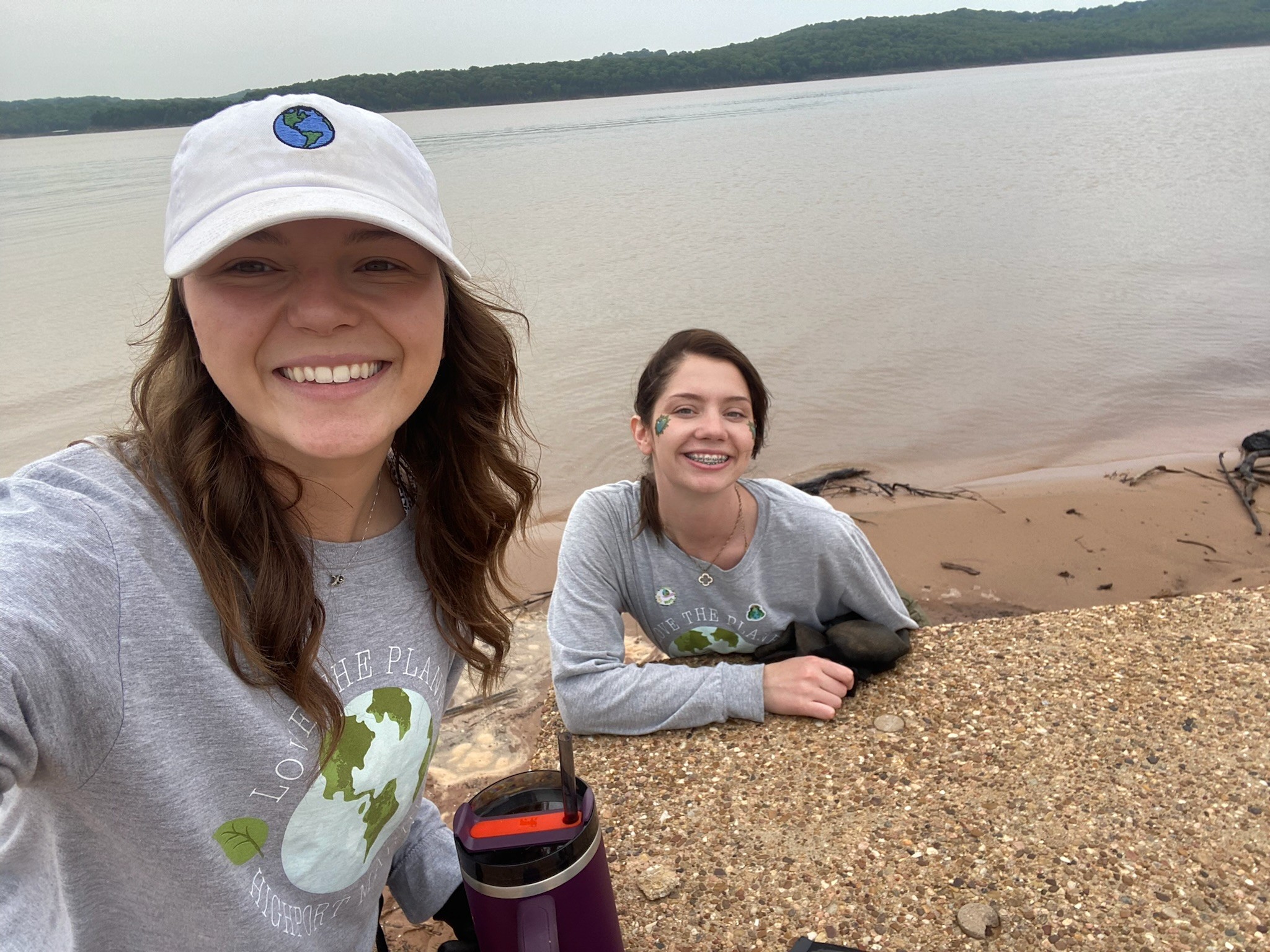 Two people wearing matching earth-themed shirts and a hat with a globe logo are enjoying a day by the lake, with one person holding a purple water bottle, under an overcast sky.