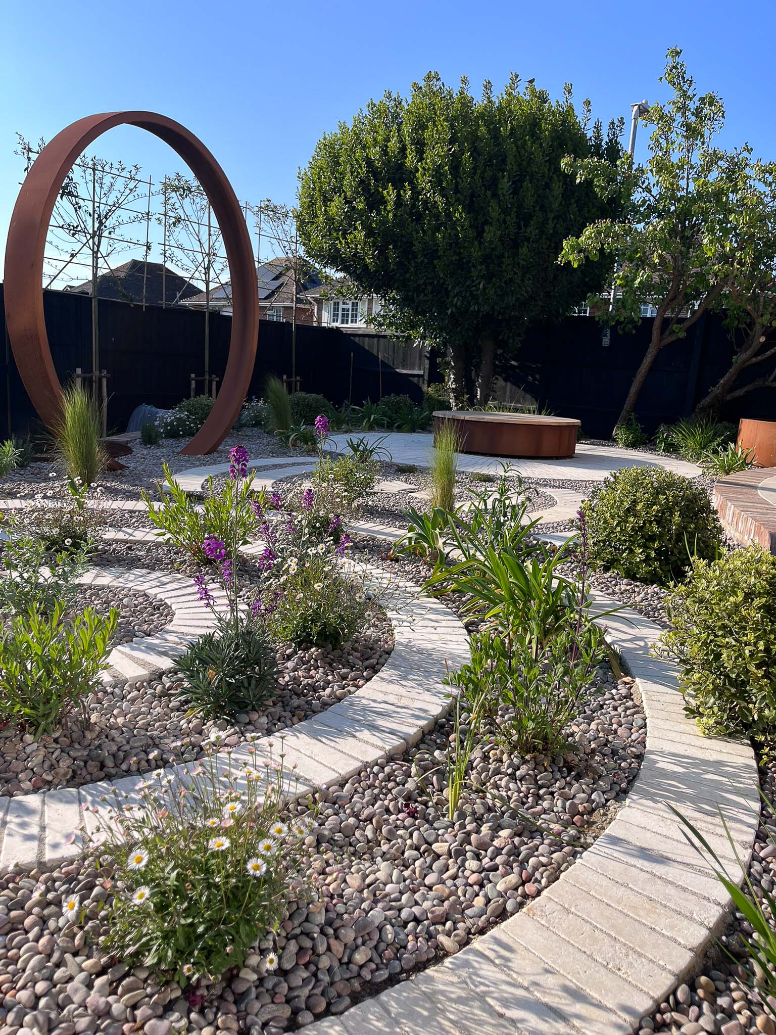 A scenic garden with curved pathways, greenery, and a circular metal sculpture under a clear blue sky.
