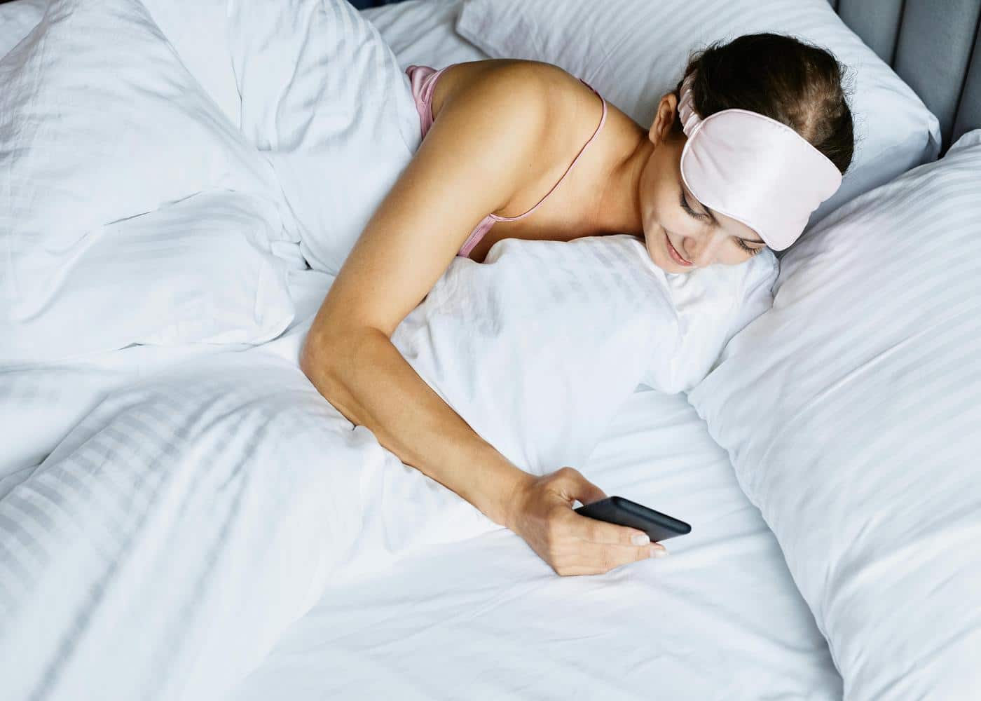 A smiling woman with eye mask on forehead laying on a bed looking at her phone