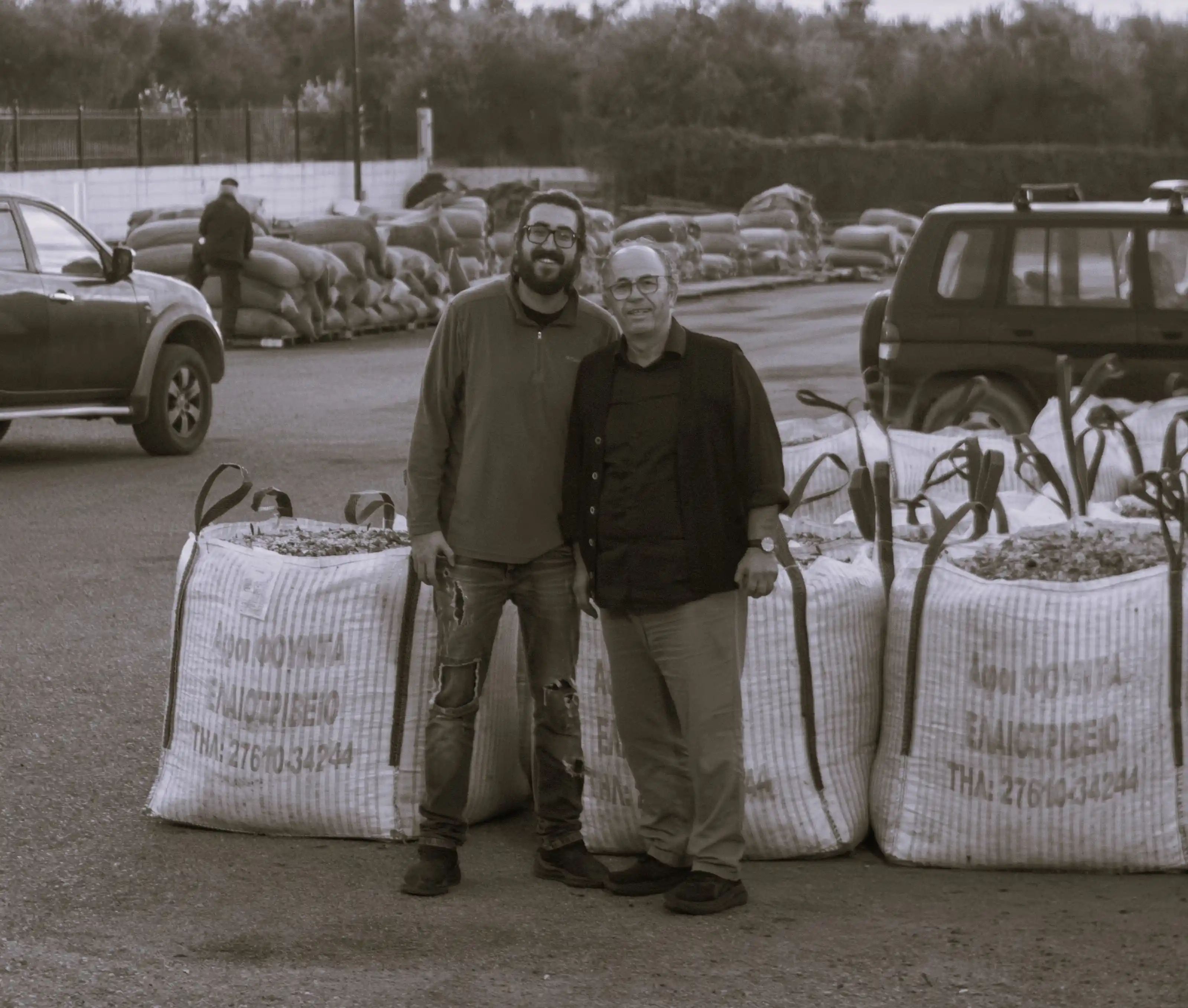 Sepia photo of two generations of the Fountas family posing behind large sacks of harvested Koroneiki variety olives at their traditional olive mill in Filiatra, Messenia.