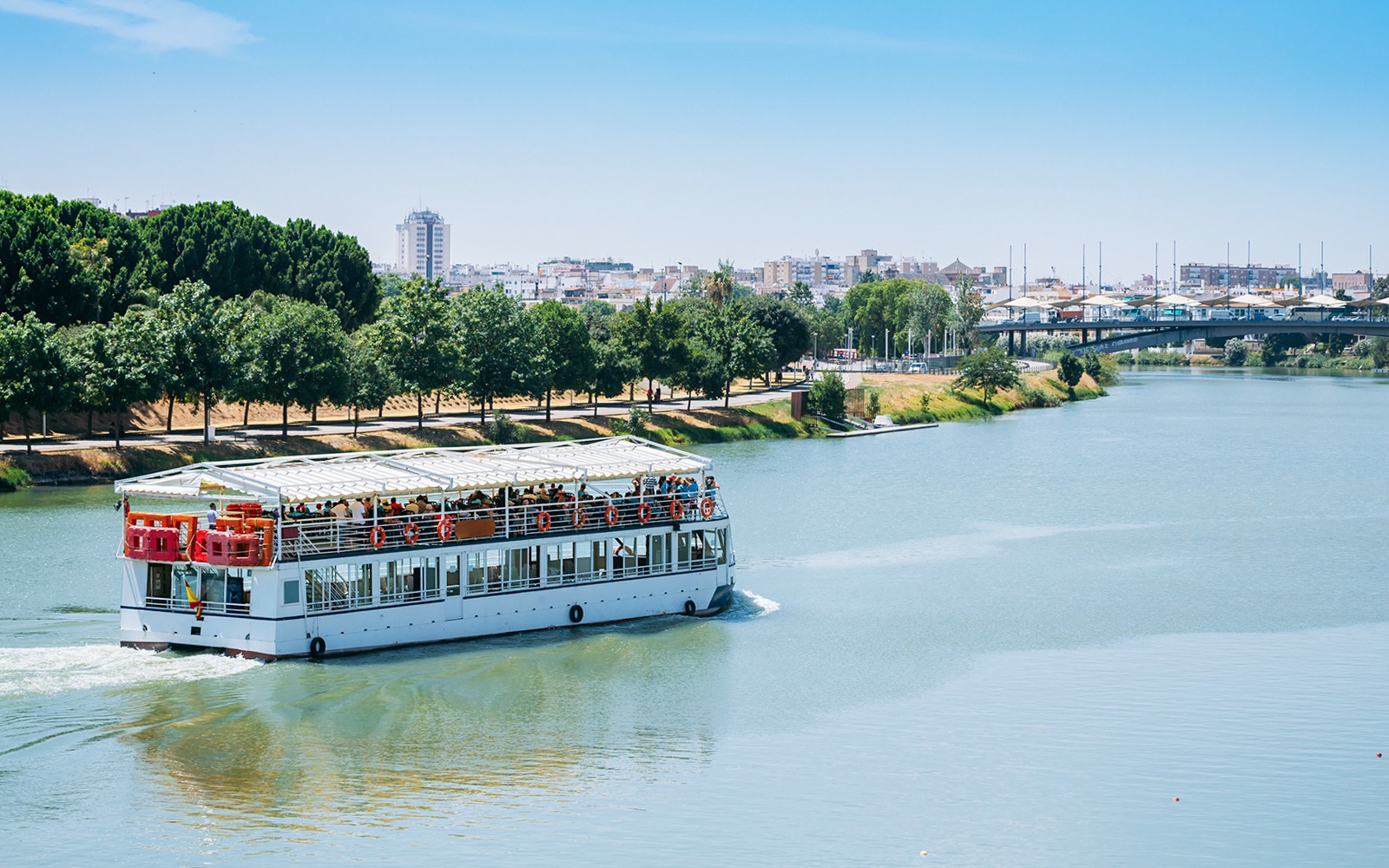 Cruise boat on Guadalquivir River in Sevilla with cityscape and trees in the background.