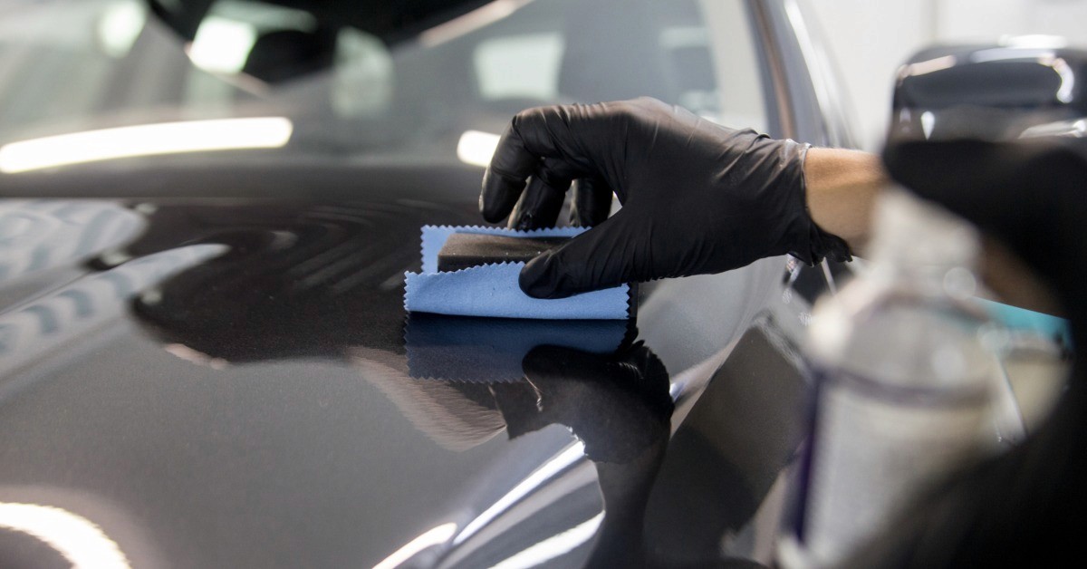 A gloved hand holding a small applicator on a dark car hood, with a blue cloth and spray bottle nearby.