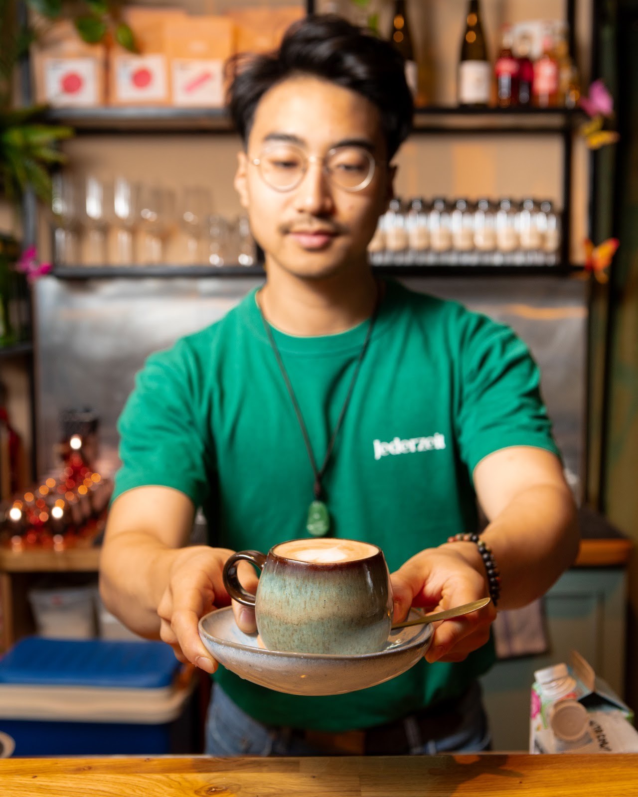 Ein lächelnder Barista in einem grünen Jederzeit-T-Shirt serviert einen Kaffee mit Latte Art in einer strukturierten blau-braunen Tasse über einer Theke.