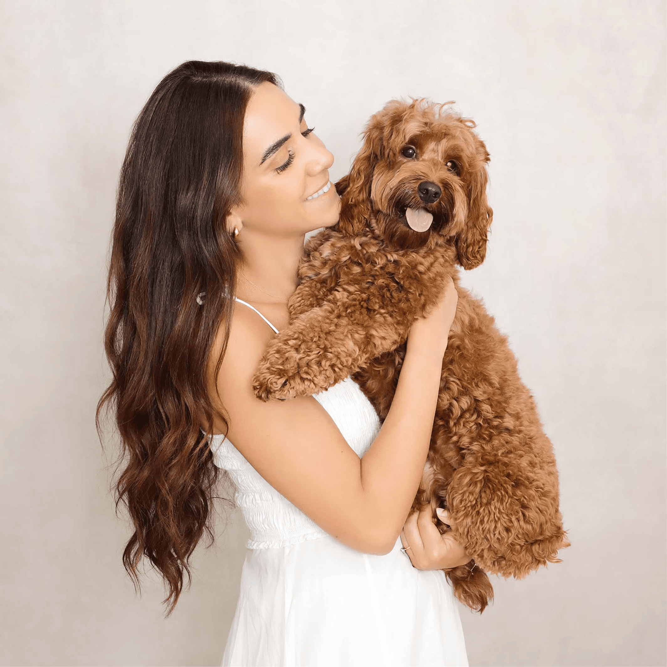 A woman in a white outfit is looking at and carrying a brown dog. The dog is sticking its tongue out. 