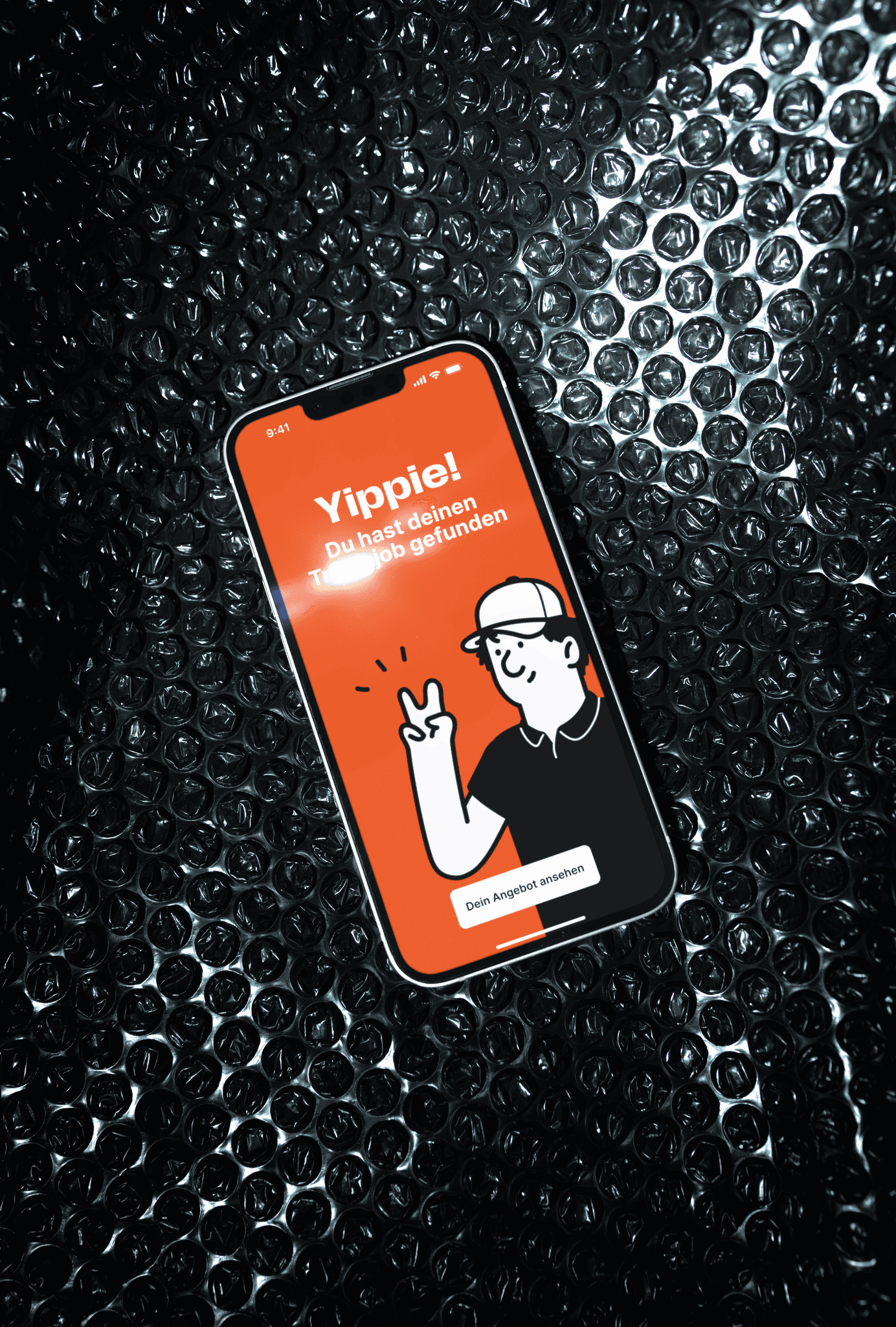 A smartphone with a cartoon character on the screen is lying on a textured bubble wrap background, featuring a red interface with the text "Yippie! Du hast deinen Job gefunden".