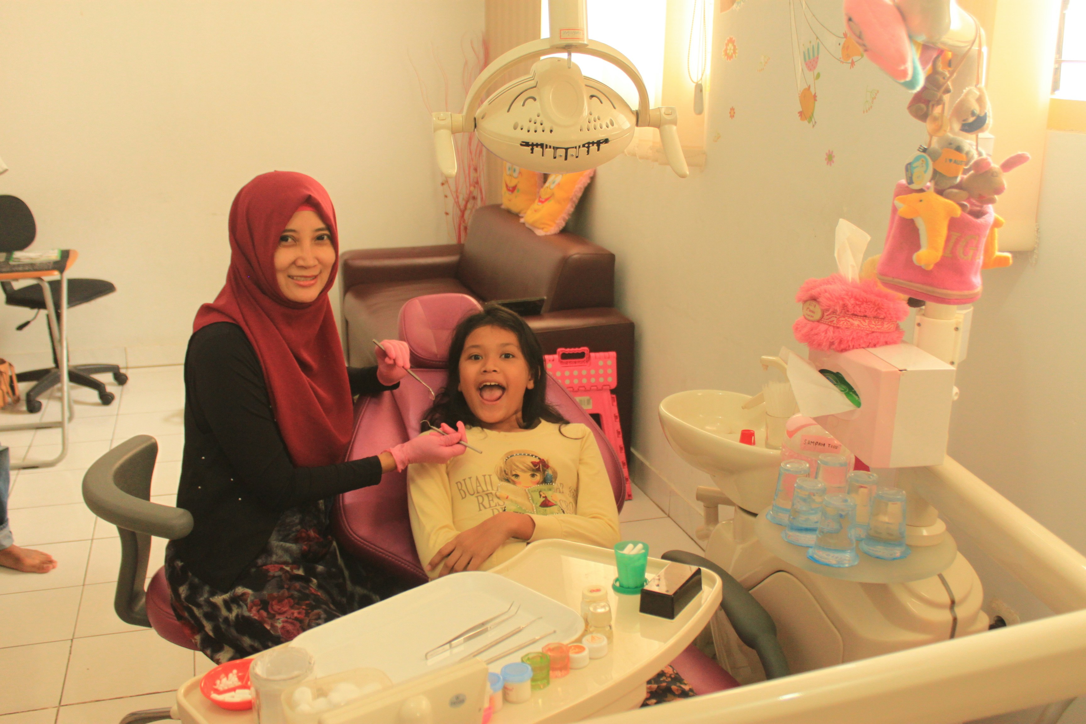 A dentist checking a young girl’s teeth while her mother sits beside her