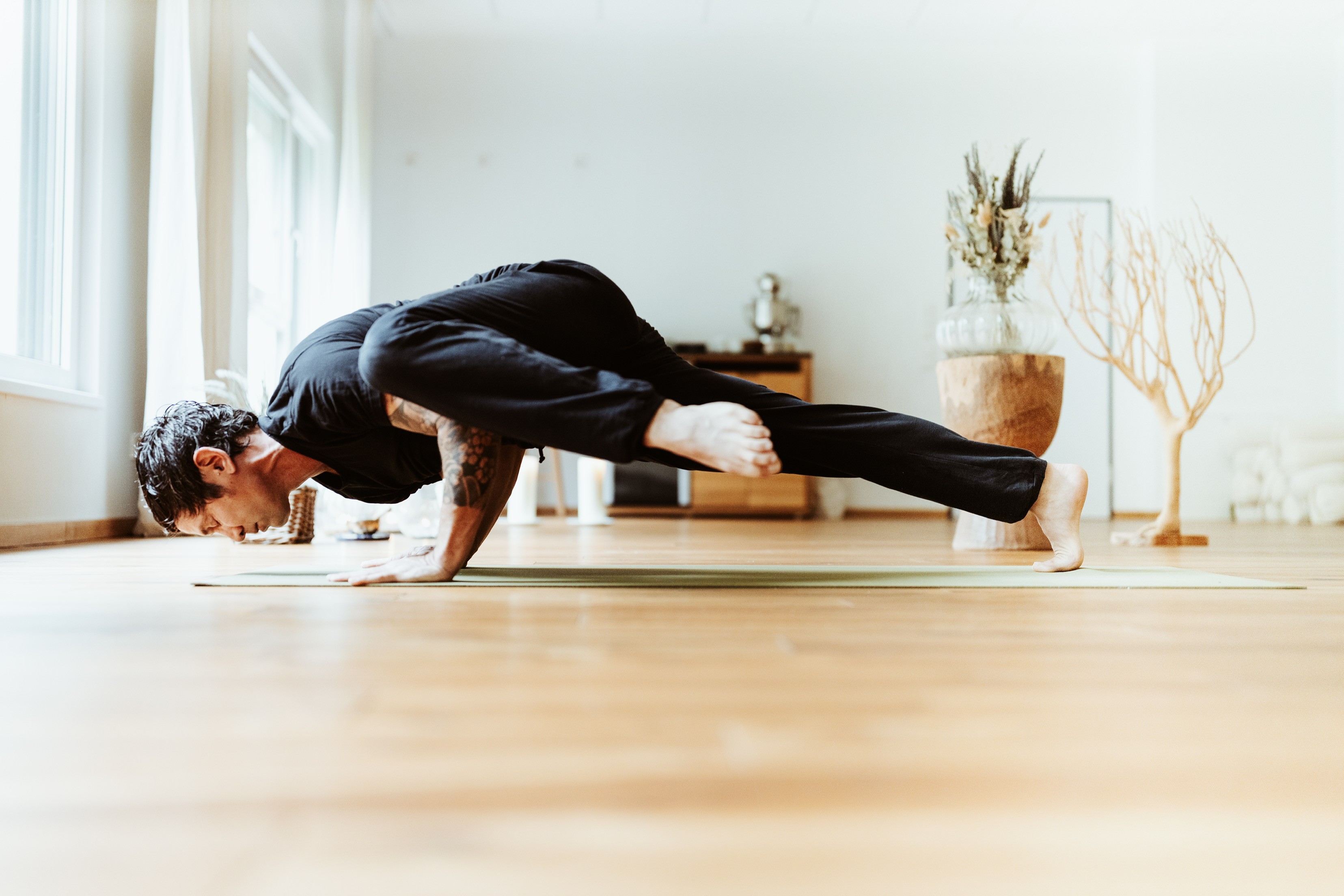A person performs a challenging yoga pose on a mat in a bright, spacious room with wooden floors and a large window, showcasing balance and strength.