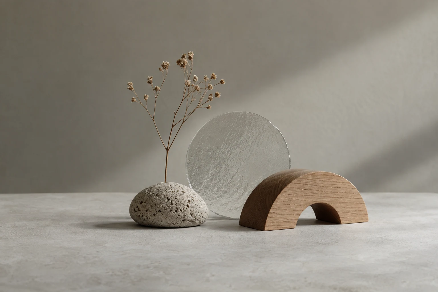 Small dry floral sprig rising from a porous pebble-shaped base, one translucent glass disk and a short arch-shaped oak object, pale gray tabletop