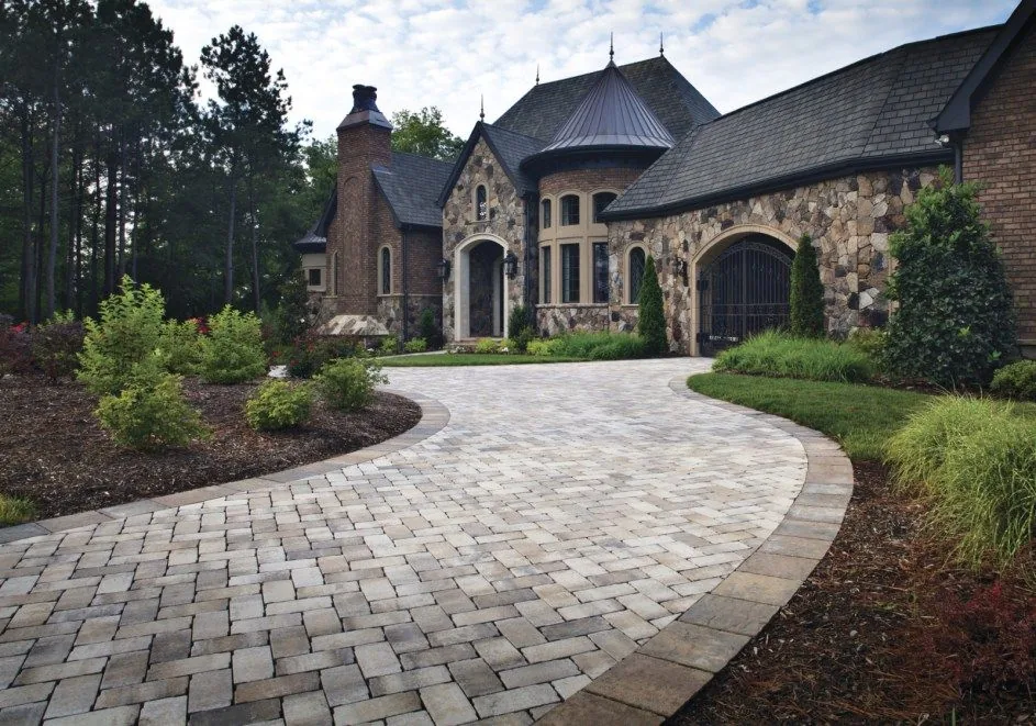 Curved paver driveway framed by planted borders in front of a stone-faced home.