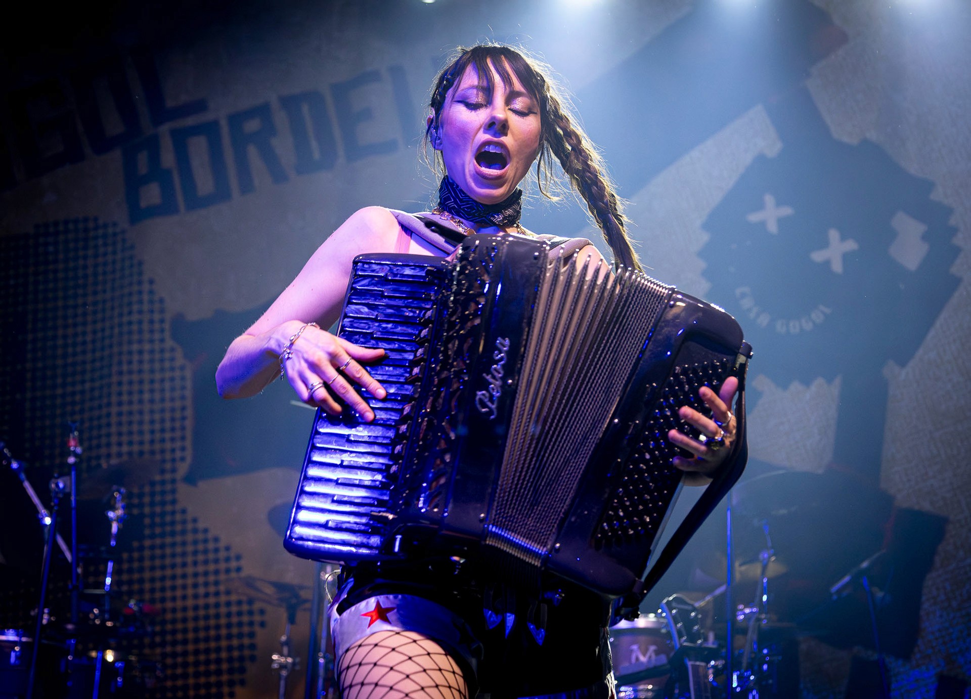 Dynamic live performance photograph of a female musician energetically singing and playing a large accordion on a darkly lit stage with blue atmosphere.