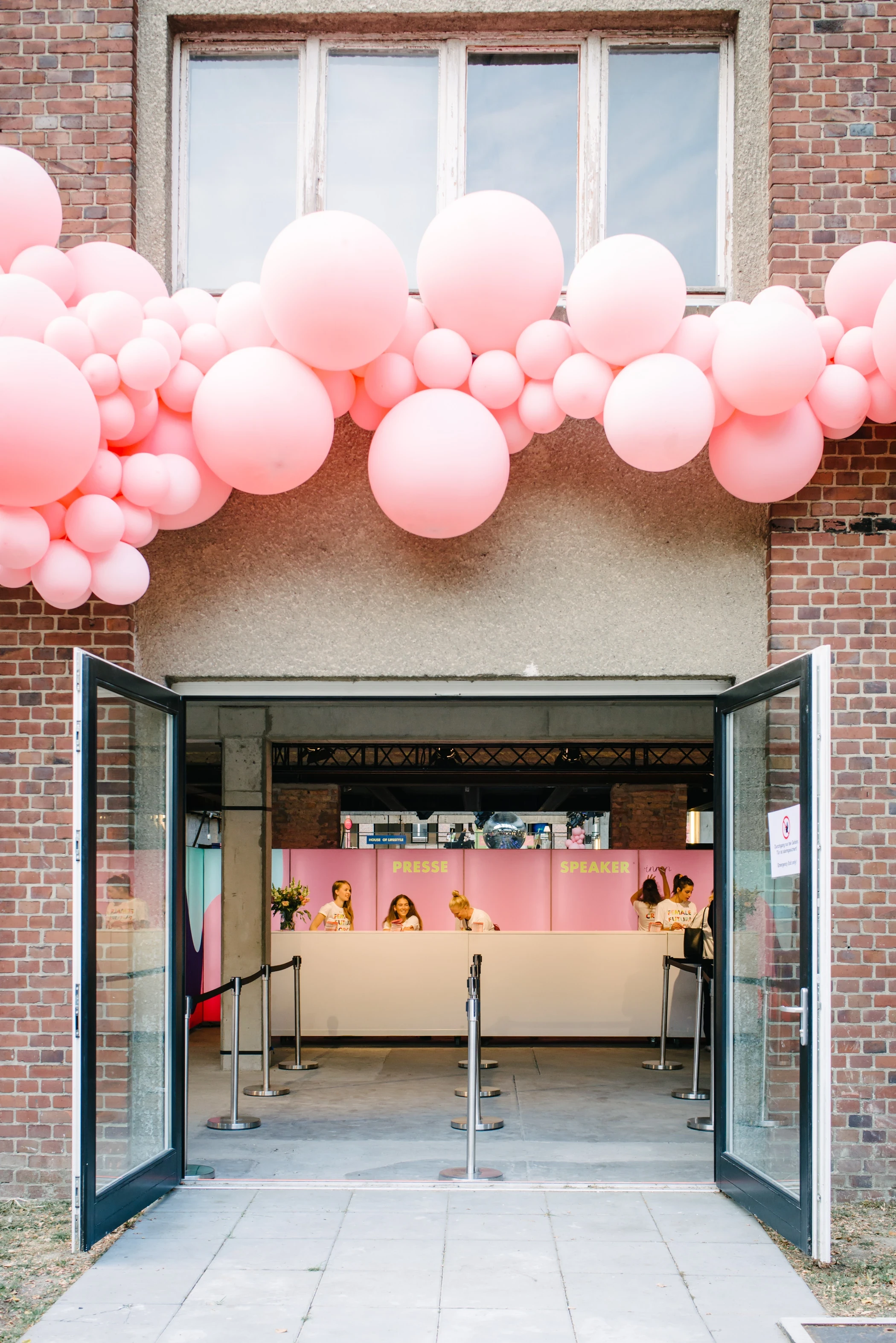 Entrance of the FEMALE FUTURE FORCE DAY with a garland of pink balloons over the door of a brick building and a pink registration counter inside.