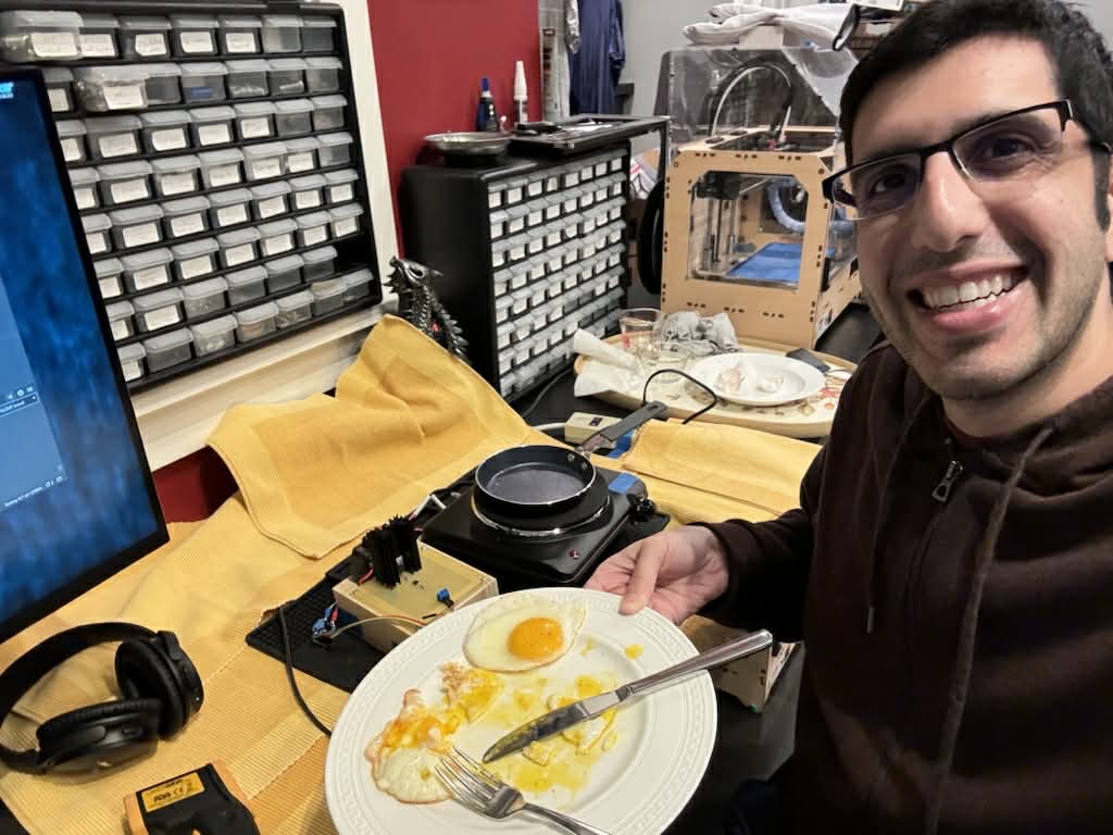 Founder Arjun Mehta sits in his workshop testing an electronics circuit board and a frying pan for cooking eggs. Equipment can be seen in the background, including a 3D printer.