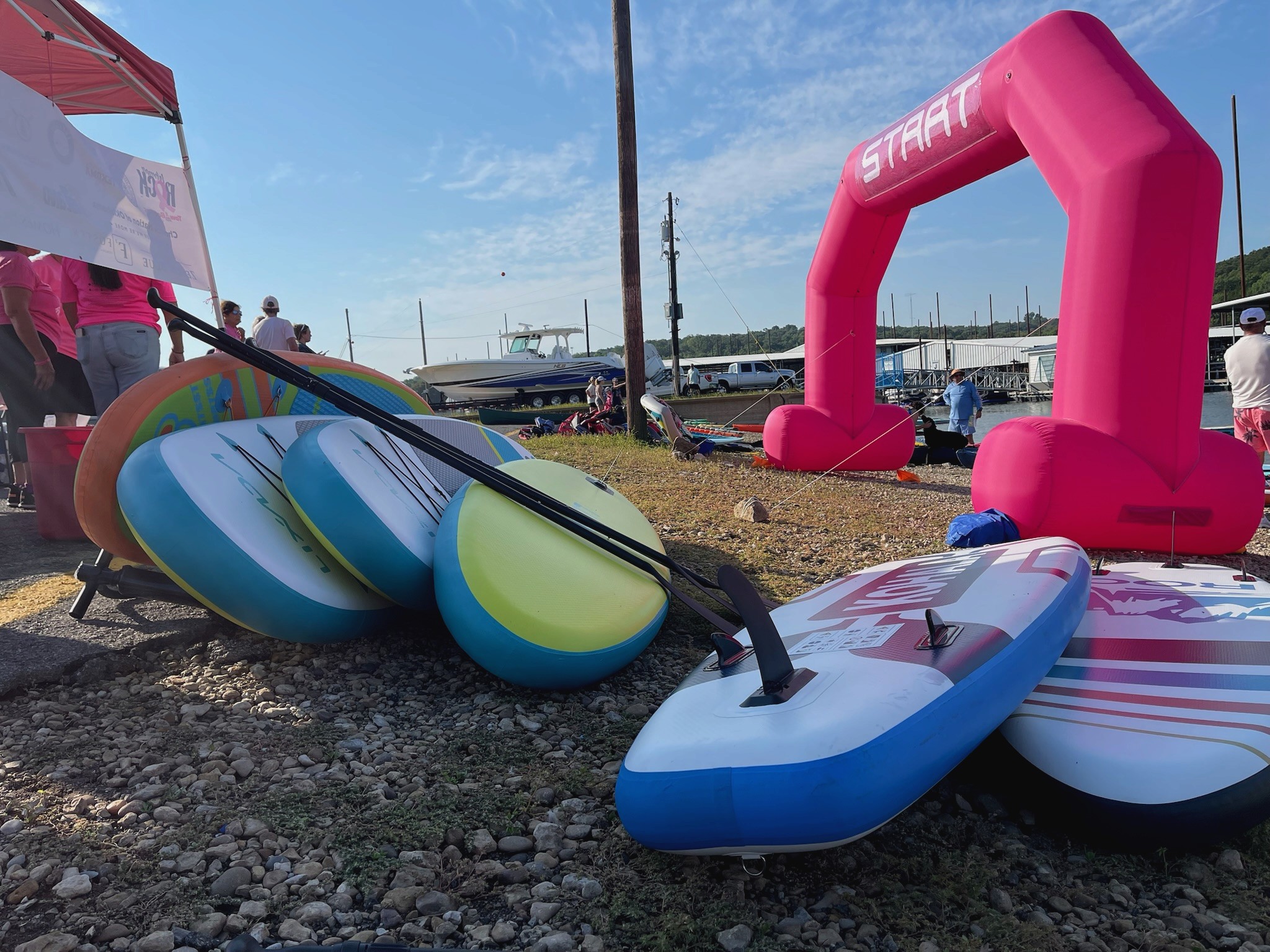 A collection of colorful stand-up paddleboards with paddles rests on a rocky shoreline beside a vibrant pink inflatable starting arch, set against a backdrop of moored boats and a cloudy blue sky.