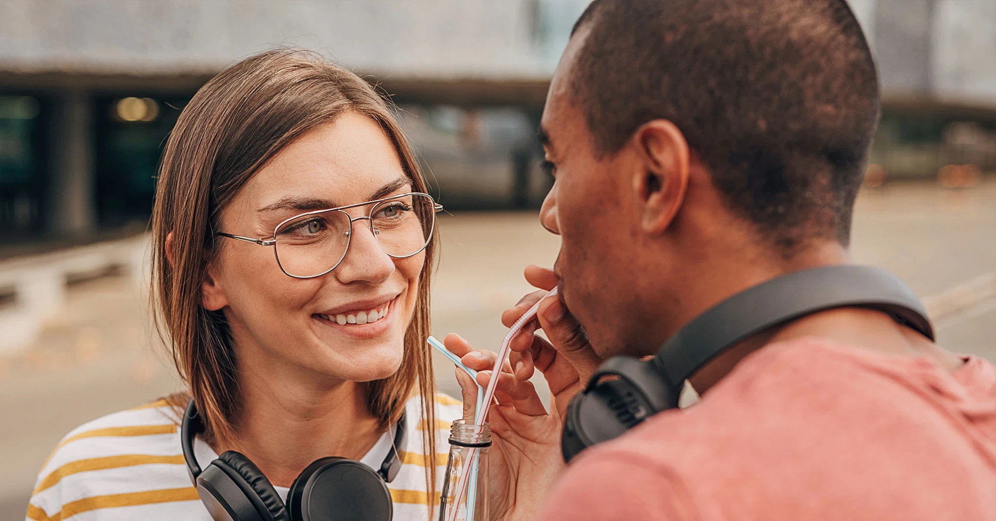  En ung, smilende kvinne med briller og en stripete topp smiler til en mann i en rød t-skjorte mens de står tett sammen og deler en flaske cola i et romantisk øyeblikk.
