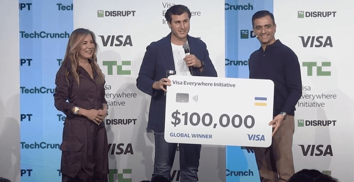 Three people stand on stage holding a large check, with logos and branding in the background.