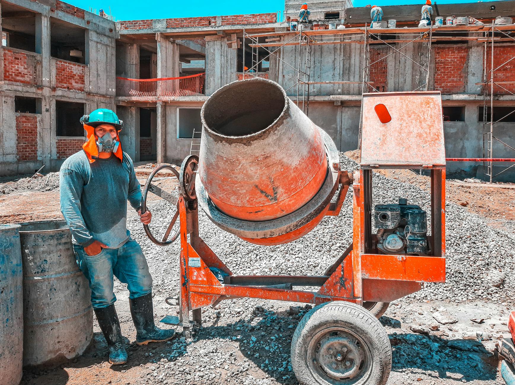 Construction worker in a green hard hat and mask stands beside an orange cement mixer at a building site