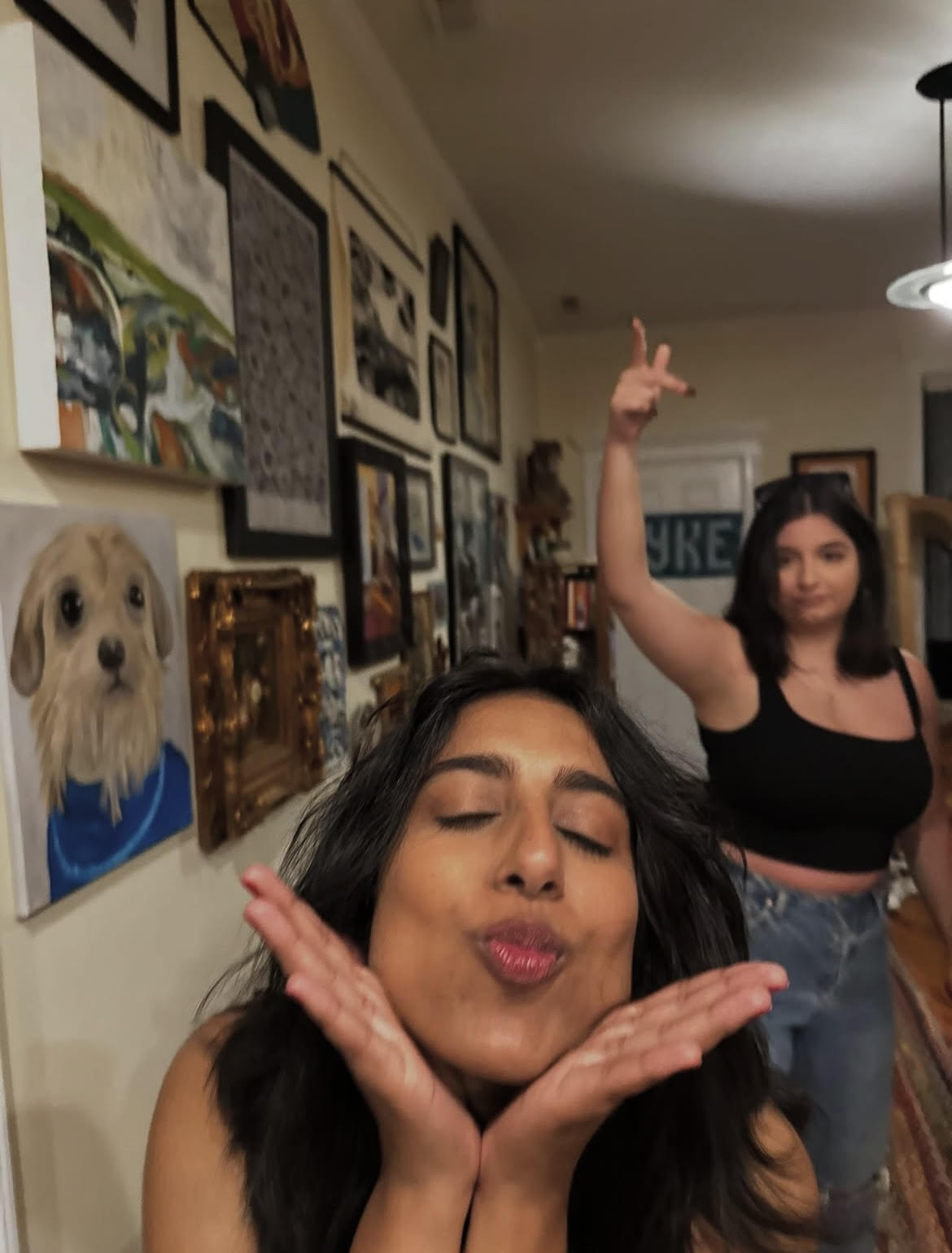 Two women are posing playfully in a cozy room filled with framed pictures on the walls. One is blowing a kiss.