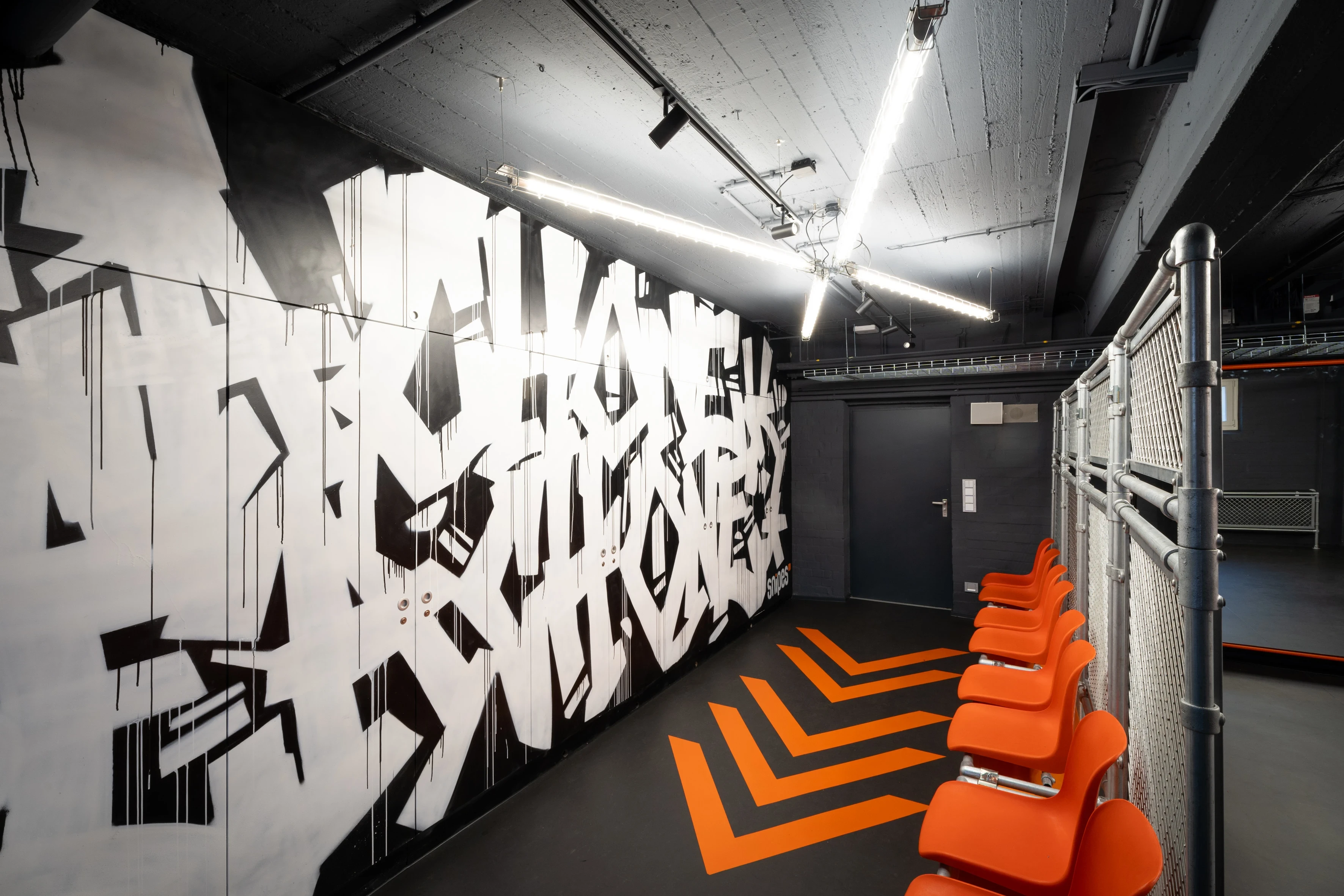 Black-and-white graffiti mural at SNIPES BASEMENT opposite orange wall-mounted seats and chain-link fence, with orange chevron floor graphics.