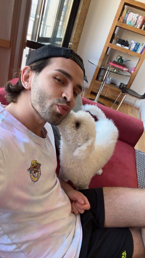 A man wearing a cap and casual clothing sits on a red sofa, making a playful pout face as a fluffy white dog snuggles close to him in a cozy living room setting, possibly highlighting a personal connection relevant to dog sitter services or a dog hotel.