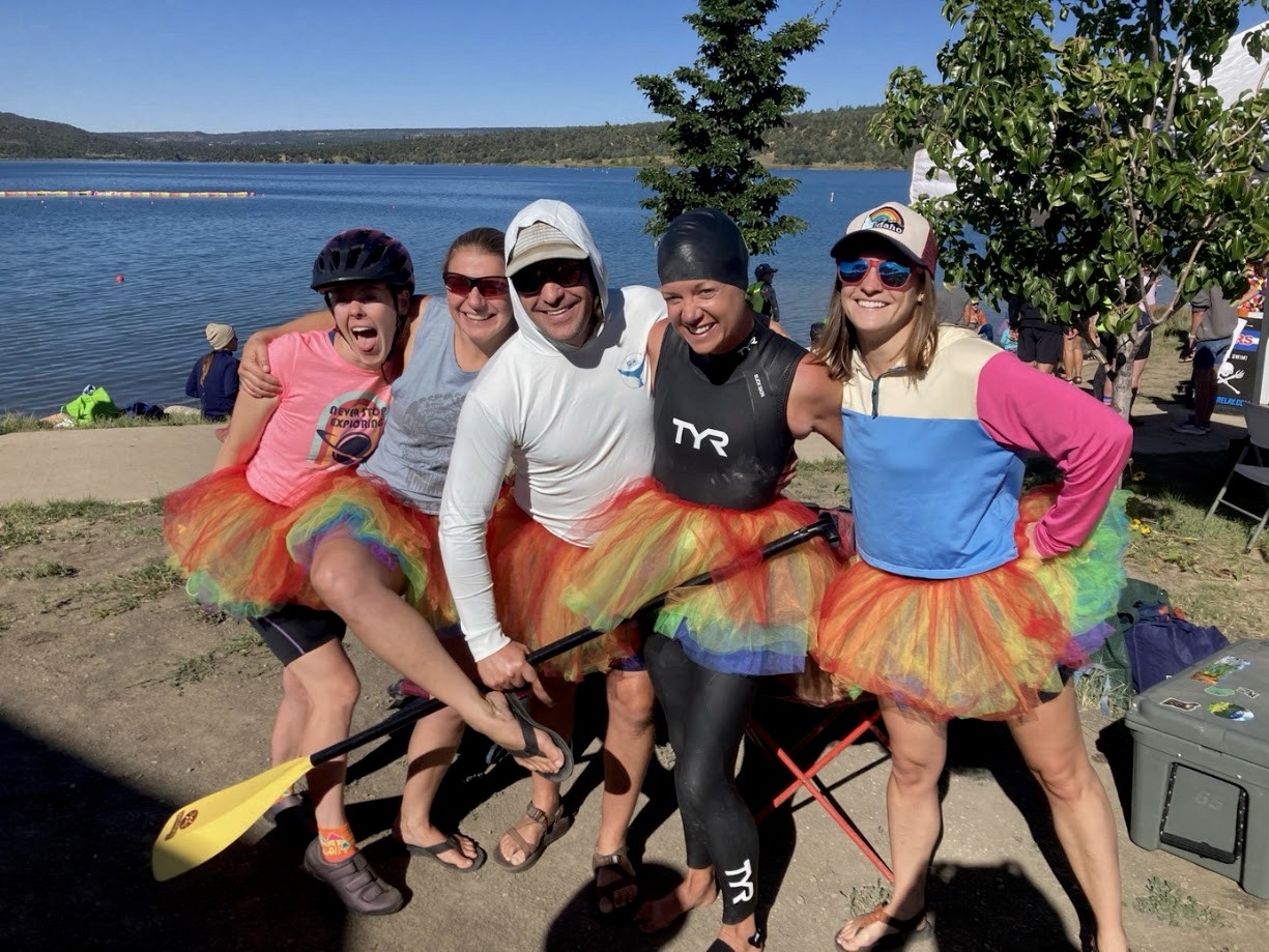 Five teammates in rainbow tutus holding a paddle at the lakeshore