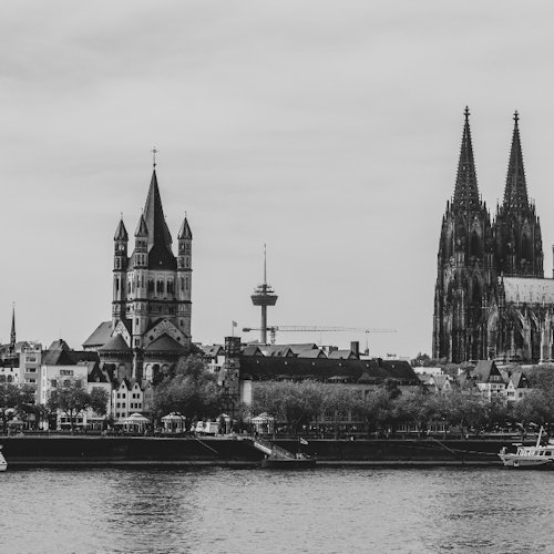 Foto hitam putih dari pemandangan kota yang menampilkan gereja-gereja besar bergaya Gotik dan Romawi di dekat sungai, dengan beberapa bangunan modern di latar belakang.