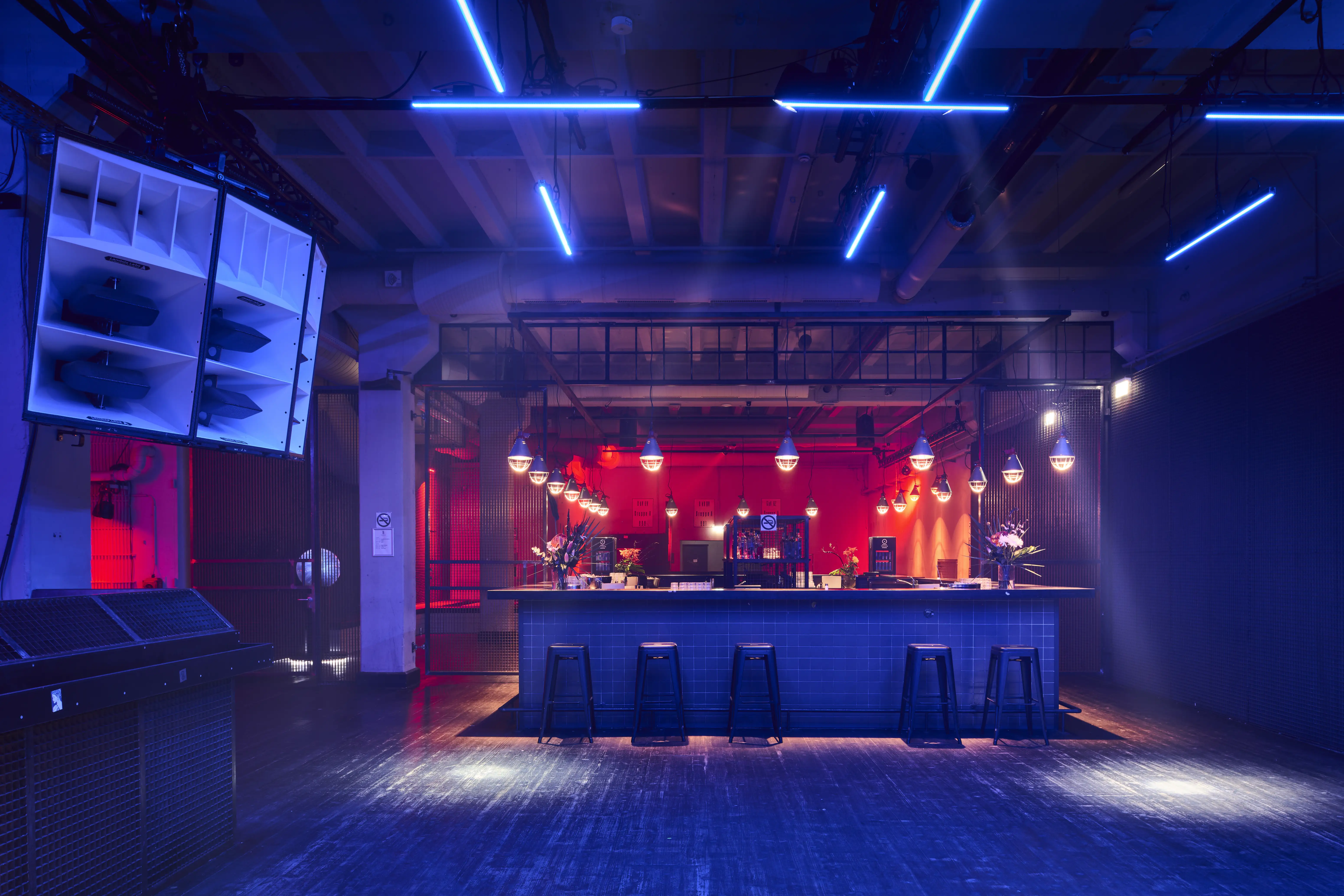 Club bar at TRESOR GLOBUS in blue light with a freestanding counter, black bar stools, caged pendant lamps, red-lit back wall and white horn speakers under blue neon tubes.