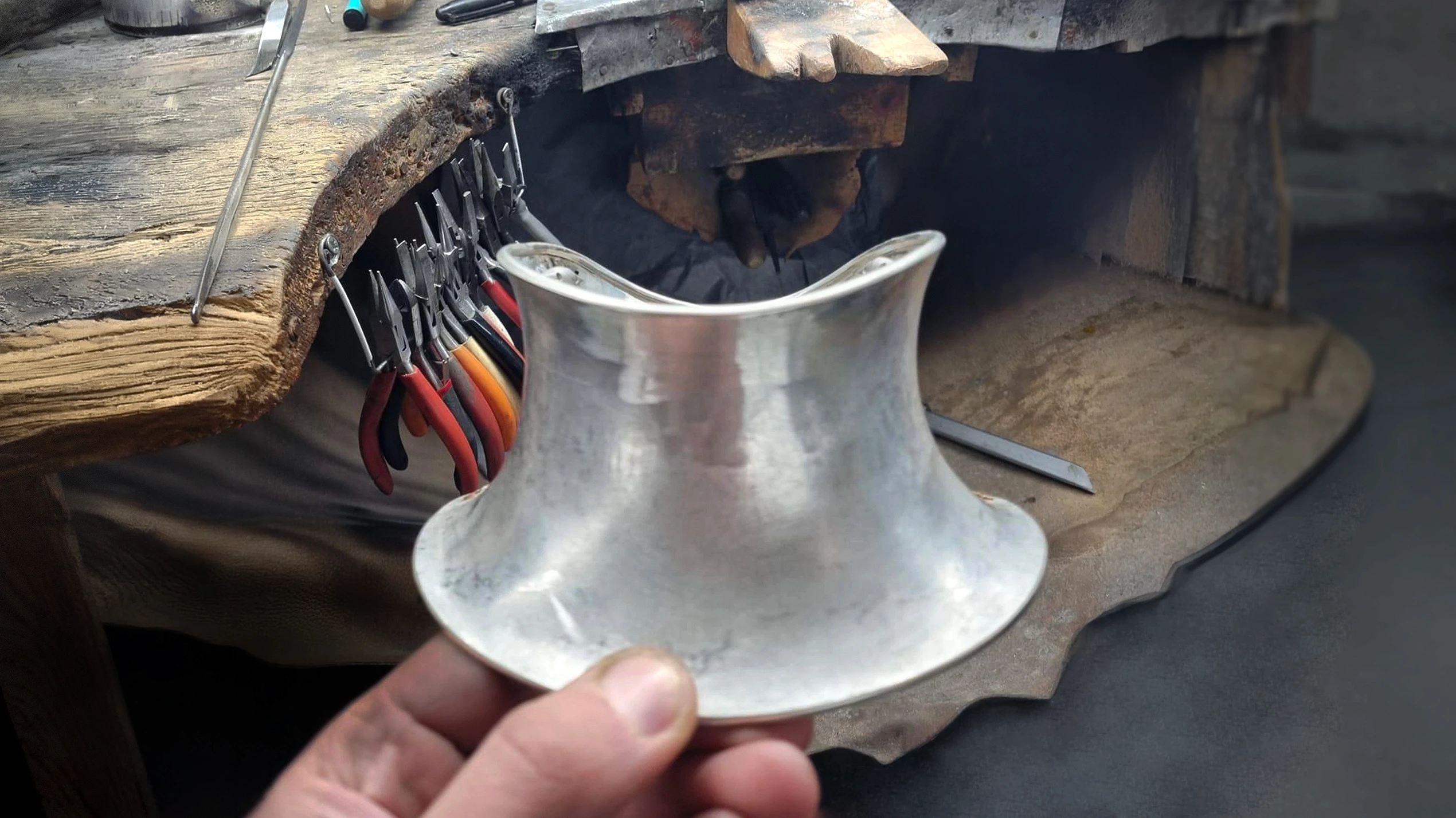 Hand holding the makings of a silver neck collar taking form, shaped around a metal mandrel on a silversmithing workbench, surrounded by workshop tools