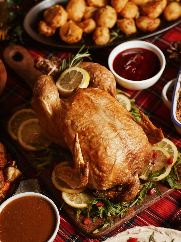 Roasted turkey garnished with lemon slices, surrounded by various festive sides on a tartan tablecloth.