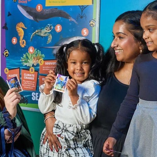 Tre bambini e una donna sorridono vicino a un colorato poster educativo sulla vita marina. Un bambino tiene in mano una carta.