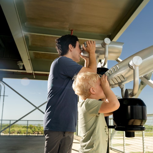 An adult and a child look through coin-operated binoculars on an observation deck with a view of a landscape in the background.