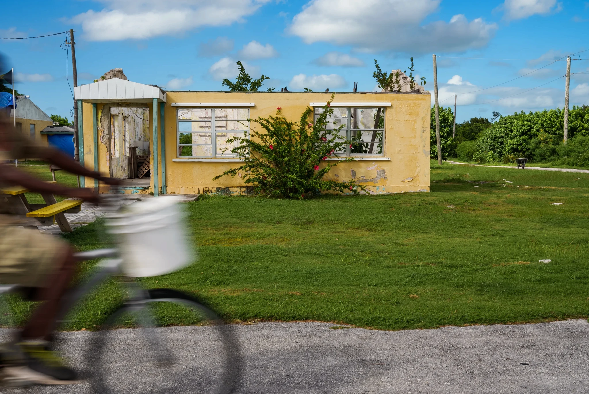 A person rides a bicycle past an abandoned yellow building with broken windows on a grassy area. The sky is blue with a few clouds, and there are electrical poles in the background.