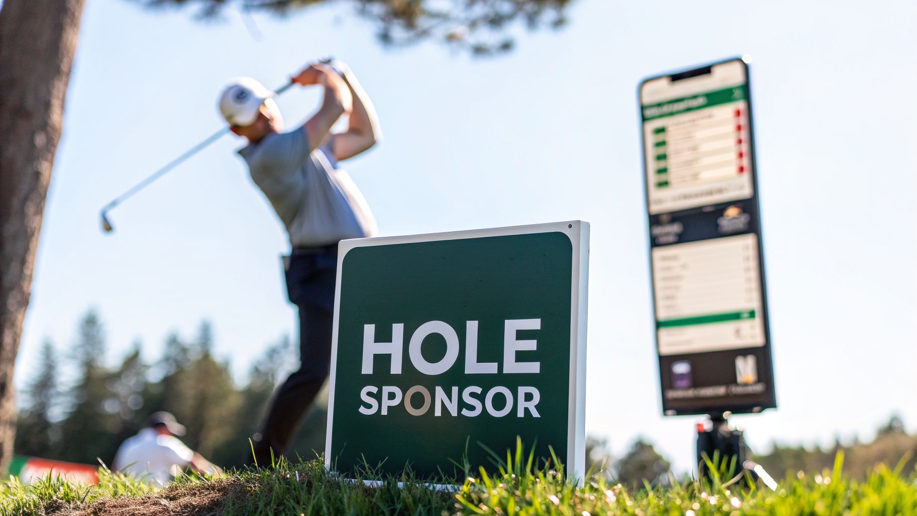 A golfer swings a club, with a 'HOLE SPONSOR' sign and digital scoreboard on a sunny golf course.