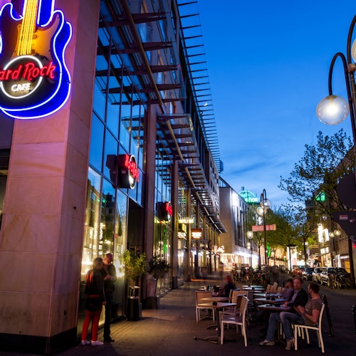 Suasana jalan sore dengan papan neon berbentuk gitar Hard Rock Cafe, meja-meja di luar ruangan, serta orang-orang yang sedang bercakap dan menikmati makan malam.