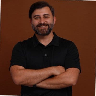 A man with a beard wearing a black shirt, looking directly at the camera with a neutral expression.
