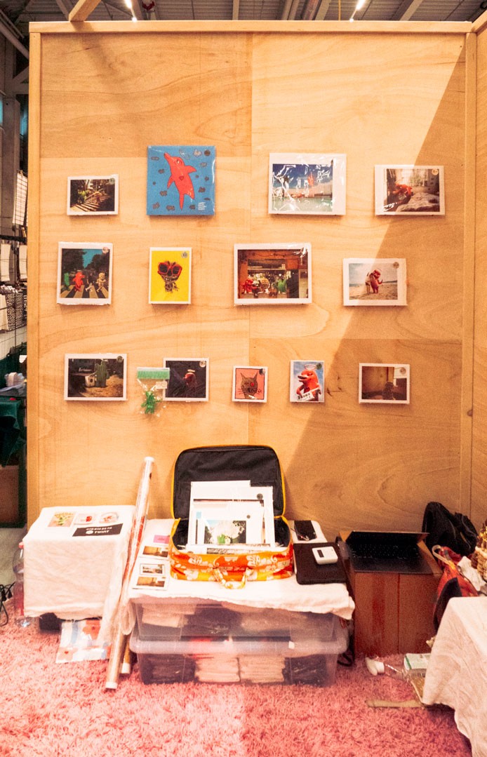 A market stall with a plywood back wall displaying a variety of framed artwork and photographs of Loveboy at the Herofest. The stall also features a table with art supplies, prints, and a suitcase.