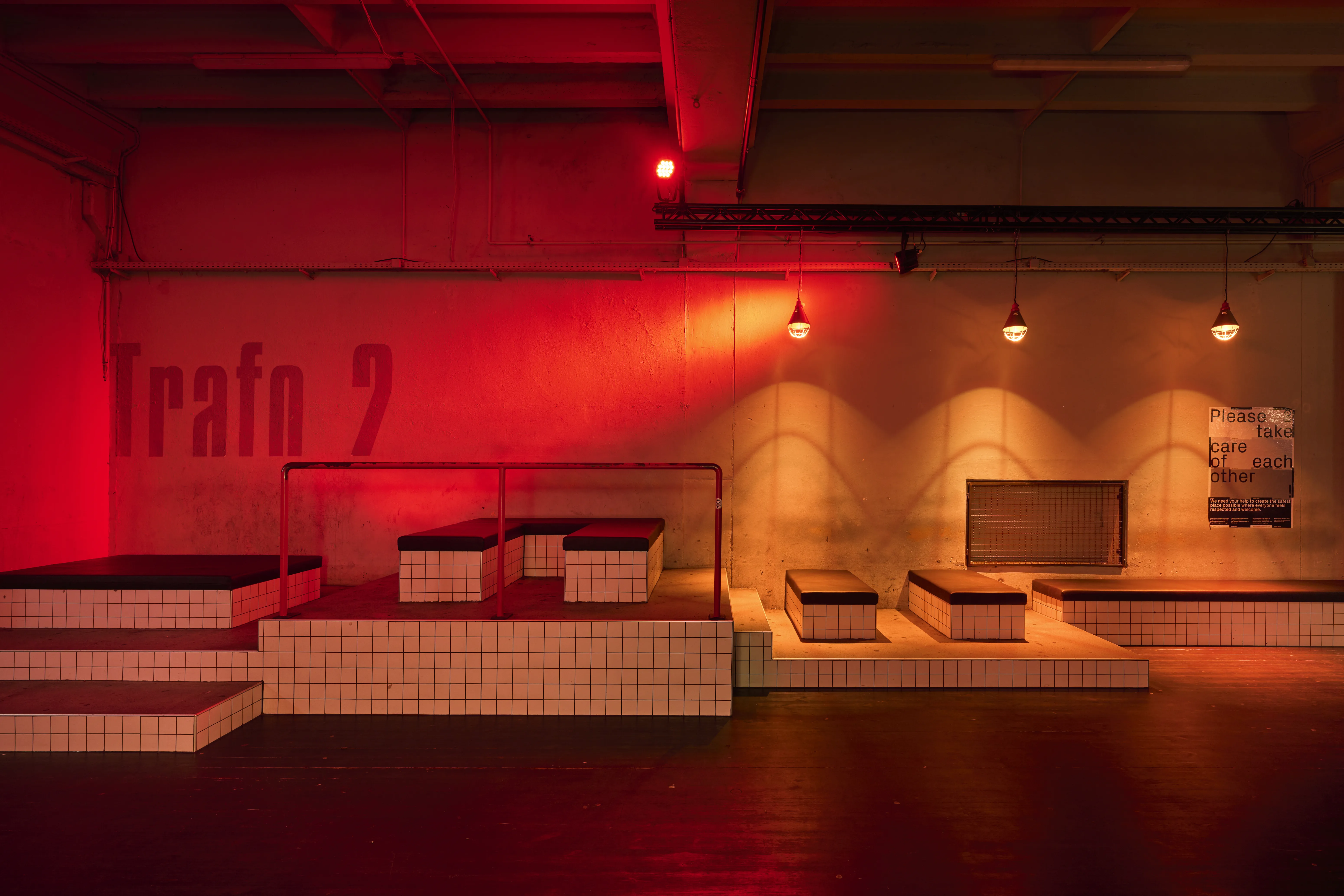 Seating landscape at TRESOR GLOBUS with white-tiled platforms and black cushions, red metal railings, warm pendant lights and the Trafo 2 lettering on a red-lit wall.