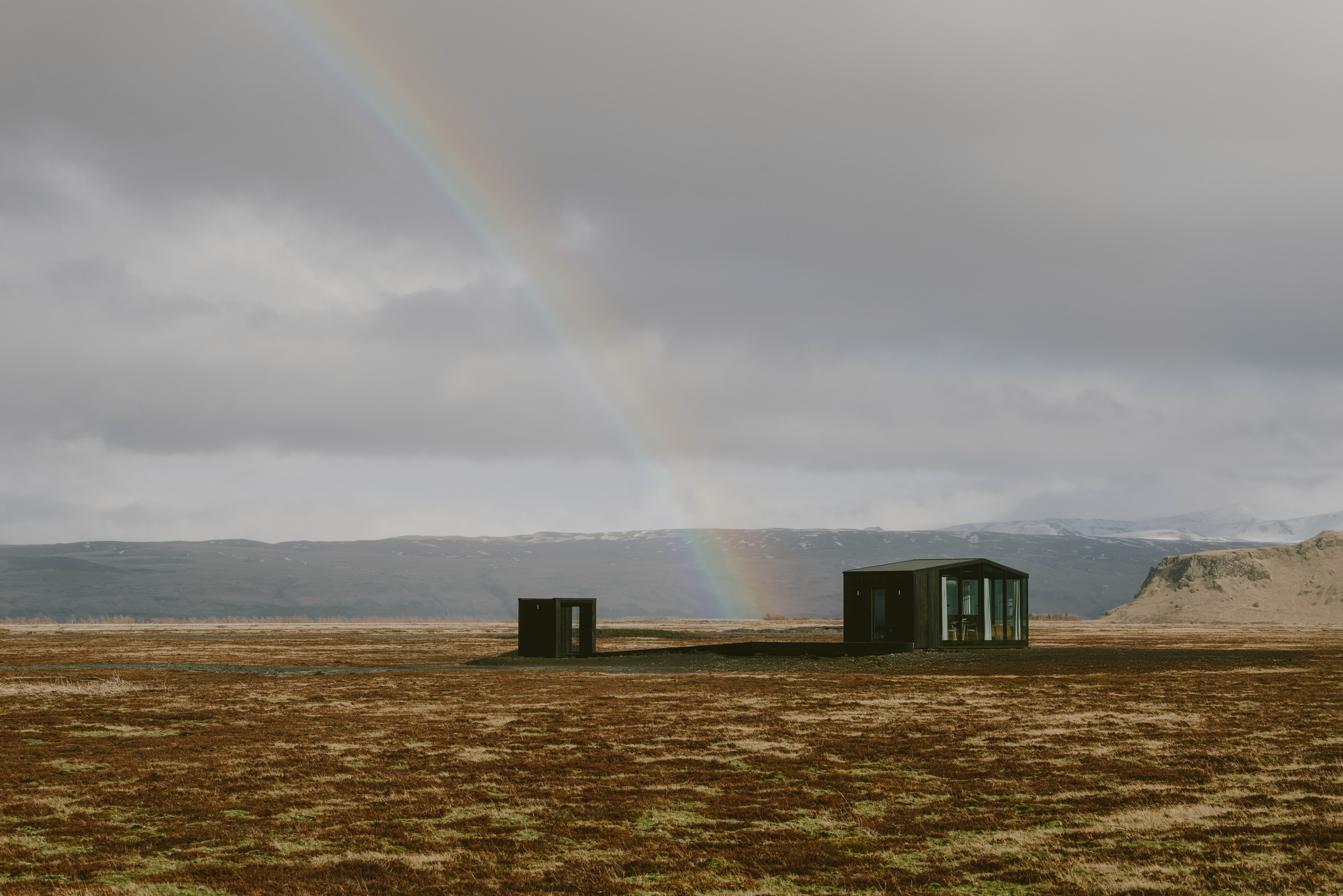 Remote modern cabin with a private sauna set in a vast, open landscape beneath dramatic skies, with a rainbow arching over snow-capped mountains, highlighting a unique design-led retreat in nature.