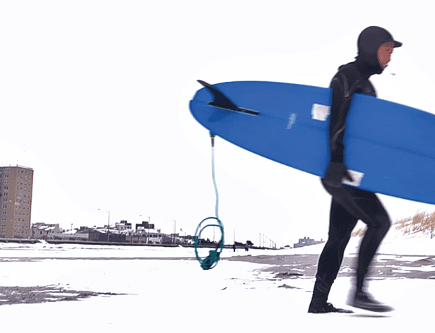 A person in a black wetsuit and hood carries a bright blue surfboard under their arm while walking across a snow-covered surface. The board has a black fin and a teal leash attached. In the background, a tall building and several structures are visible under a bright, overcast sky. The ground is covered in snow, with patches of darker ground visible near the water’s edge.